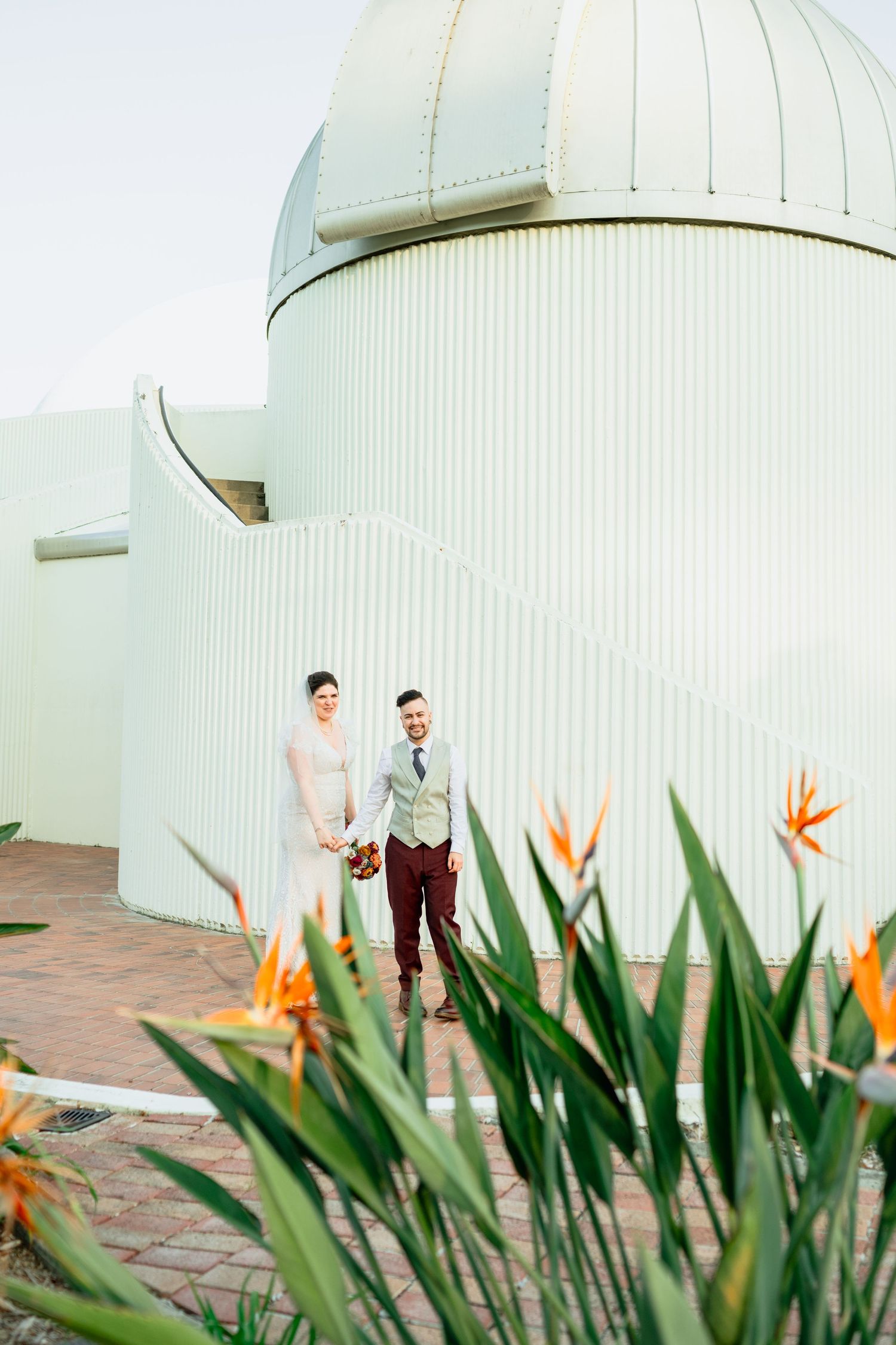 Bride and groom hold hands outside a domed observatory building with bird of paradise flowers in the foreground.