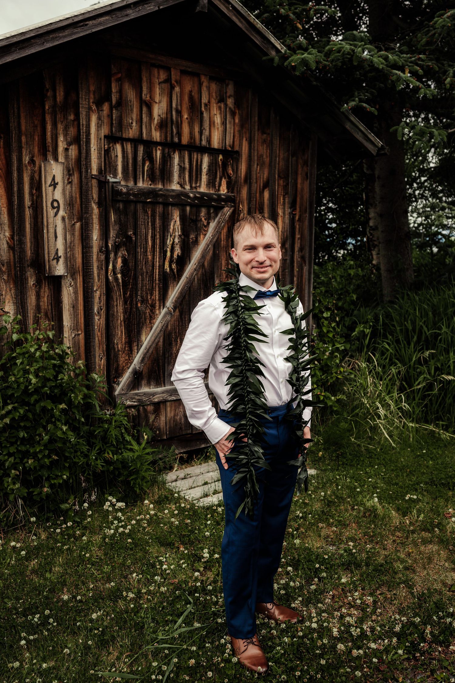 Groom in blue suit with green leaf garland smiling in front of rustic wooden barn.