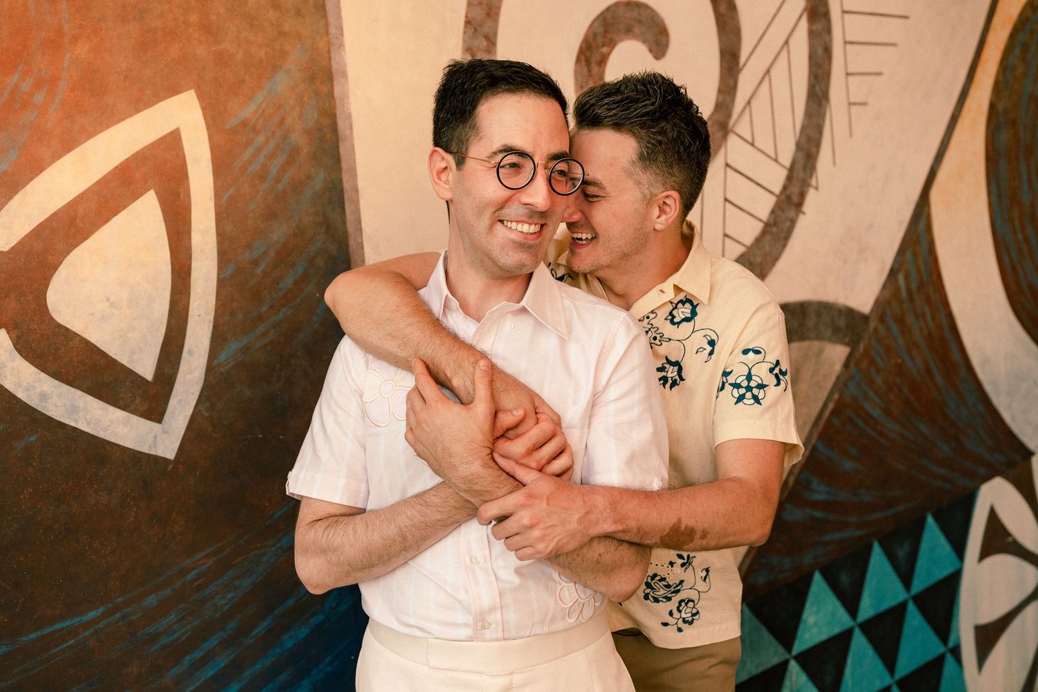 Two men embracing and smiling in front of a colorful mural, one wearing glasses and a white shirt.
