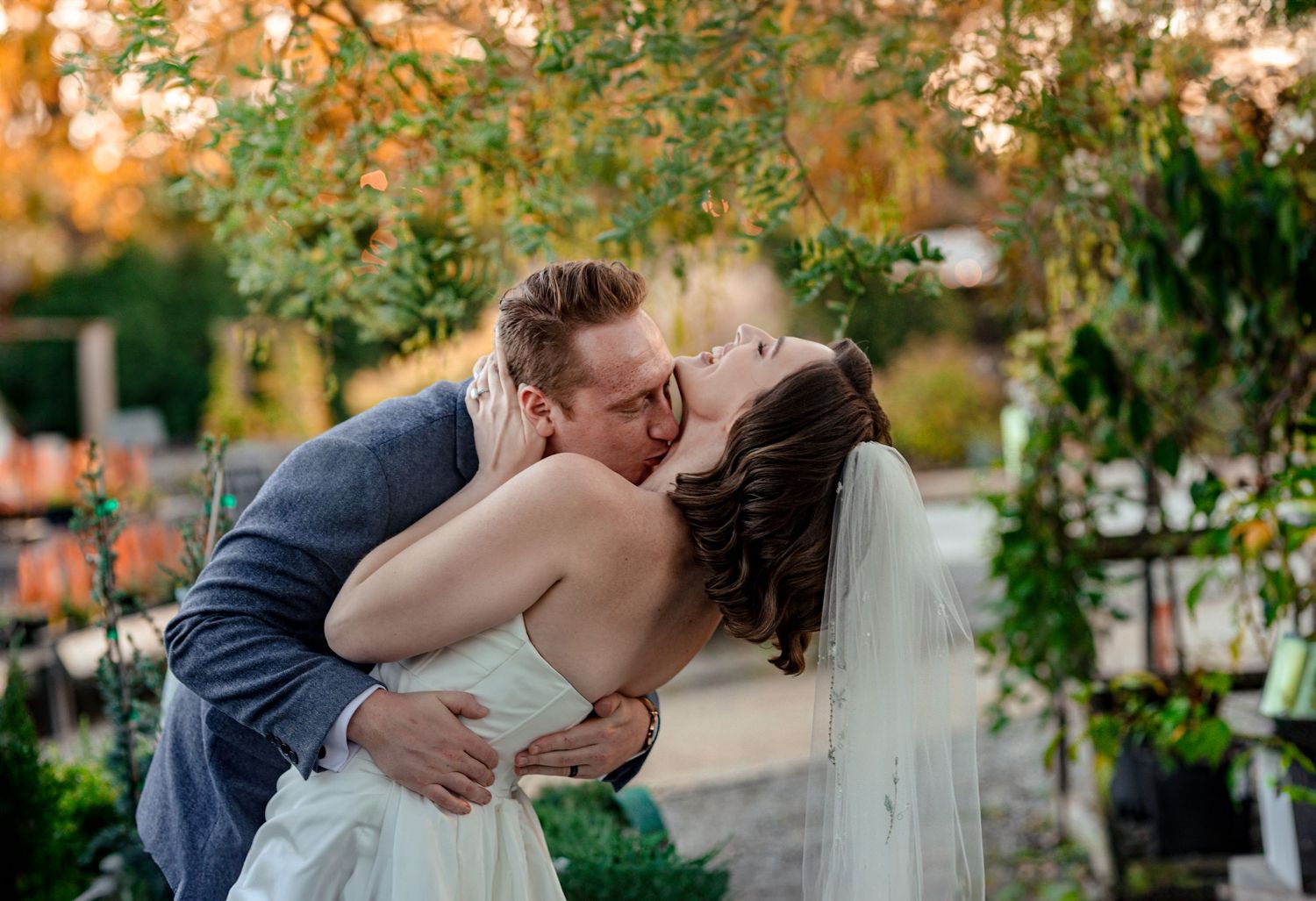 Bride and groom sharing a romantic kiss outdoors under lush green foliage during their wedding ceremony as the sun sets in fall.