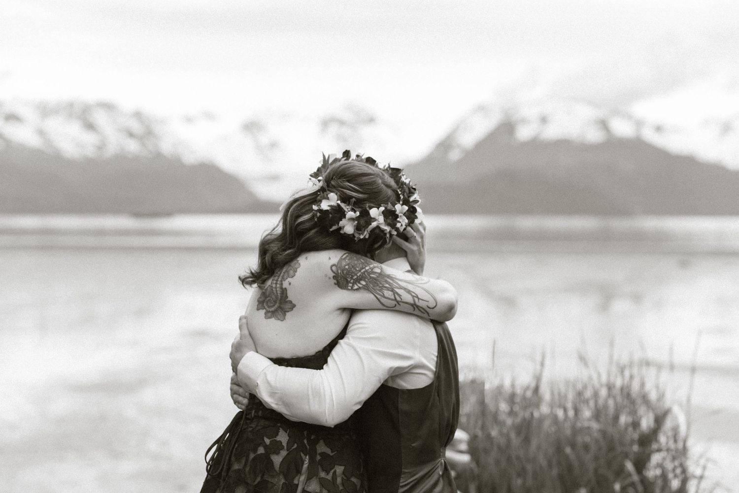 Black and white photo of couple embracing with flower crown, snowy mountains and lake in background.