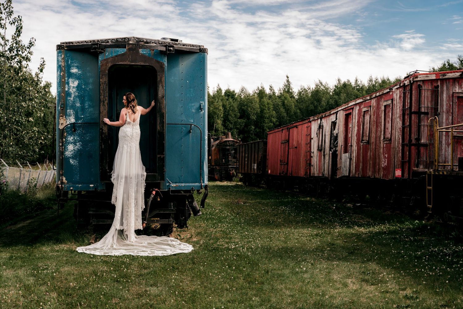 Bride in white gown standing in doorway of vintage blue train car at rustic wasilla museum of transportation.