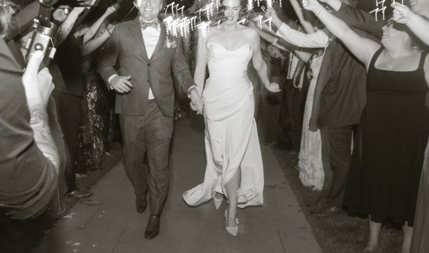 Black and white photo of a bride and groom walking through a sparkler tunnel at their wedding reception.