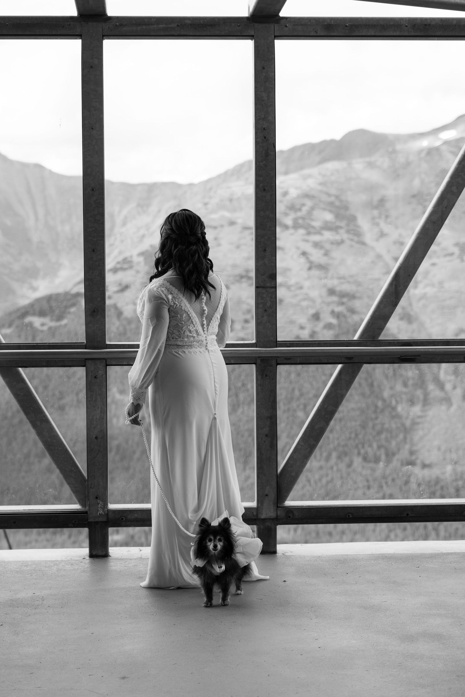 Black and white photo of a bride in a white gown holding a small dog, gazing at mountain views through large windows.