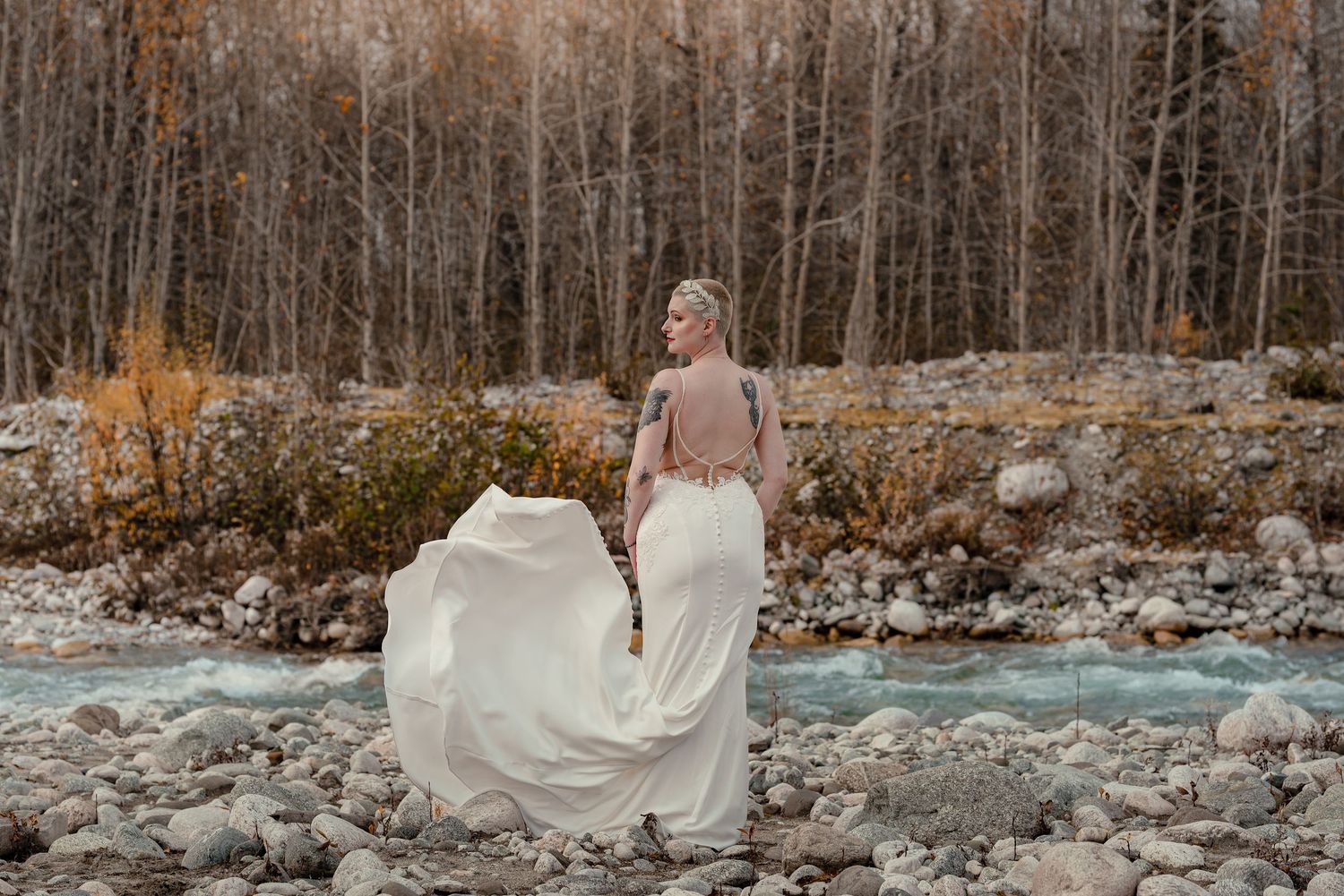 A flowing white wedding dress billows in the wind against a backdrop of winter trees and rocky terrain.