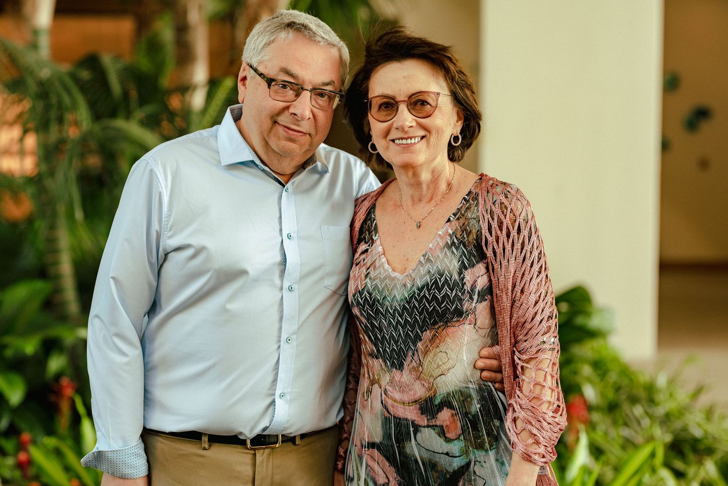 Smiling older couple posing together outdoors, surrounded by tropical greenery, woman wearing sunglasses and floral top.