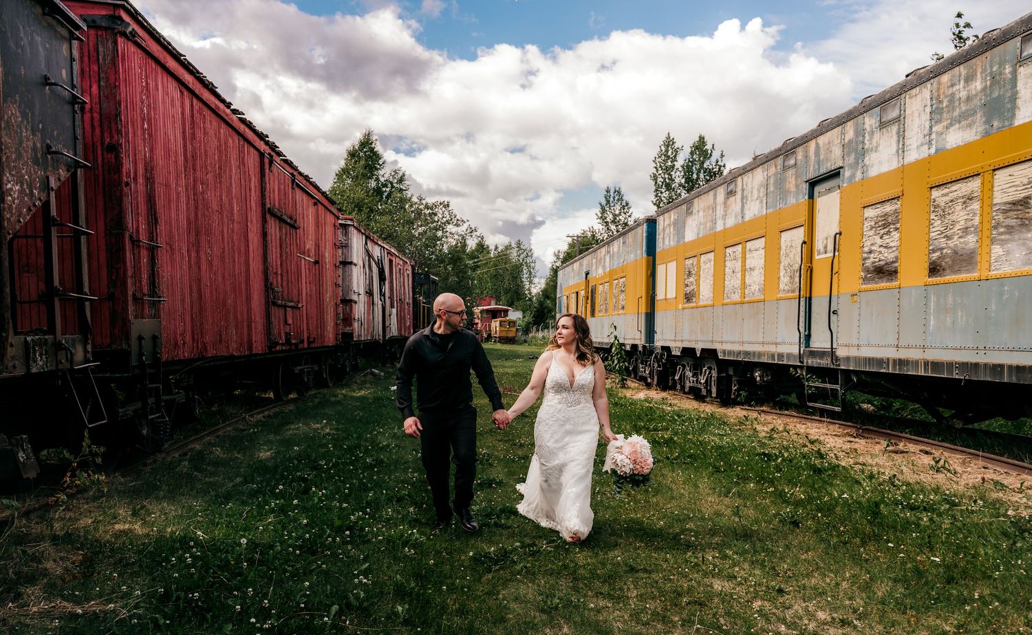 Bride and groom walk hand-in-hand between vintage train cars on a cloudy day at a scenic railway in wasilla alaska