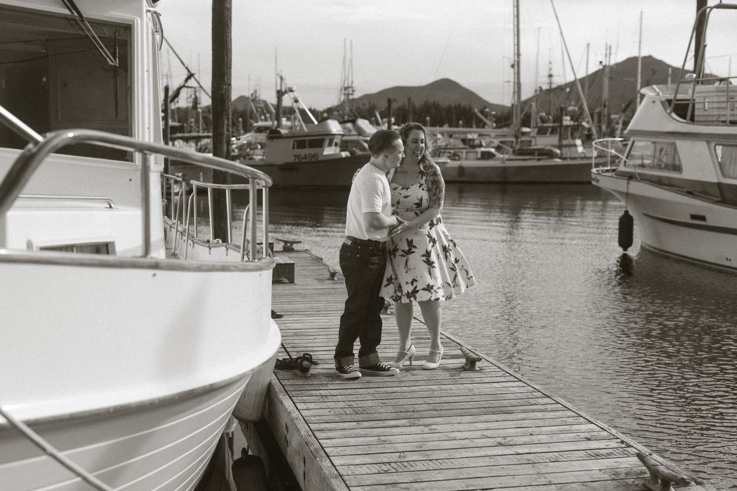 Couple standing on a wooden dock at a marina with boats and mountains in the background, black and white photo.