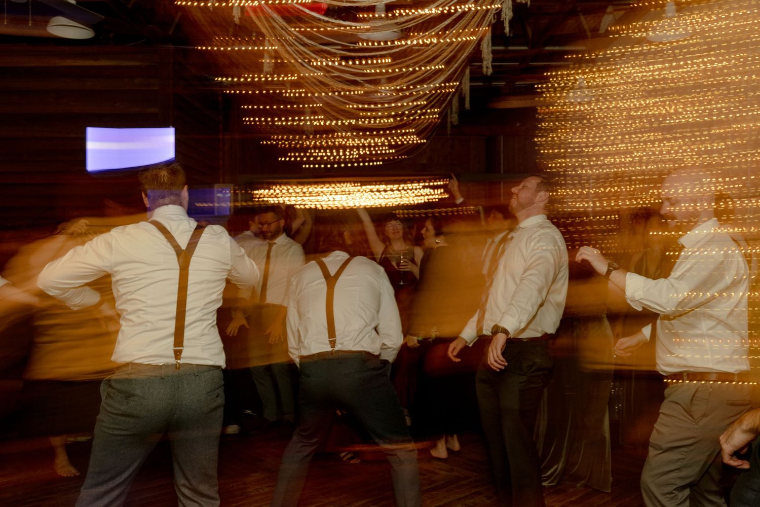 Groomsmen in white shirts and suspenders dancing energetically at a wedding reception under warm lights.