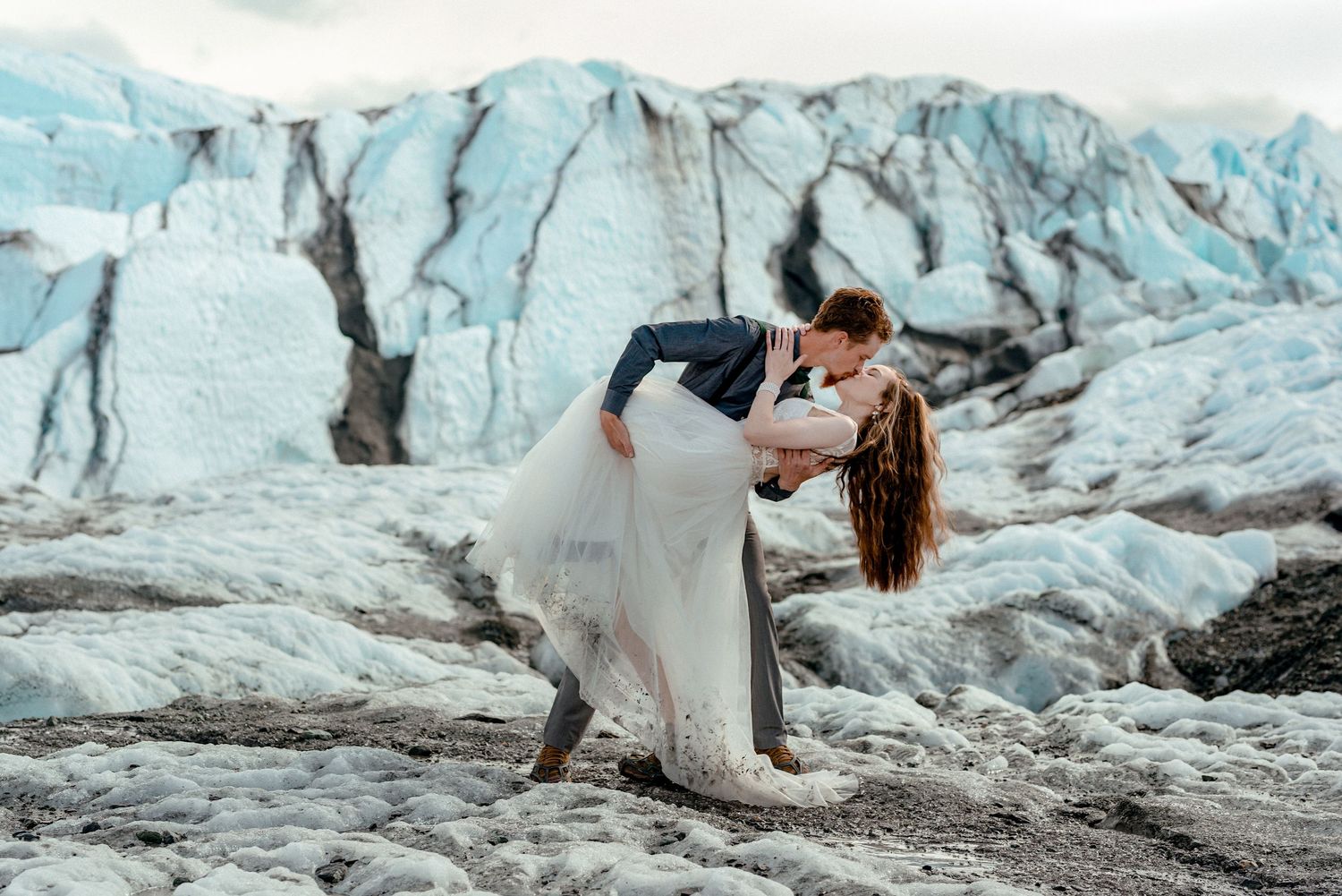 Couple sharing a romantic dip kiss in wedding attire with stunning glacier ice formations in the background.