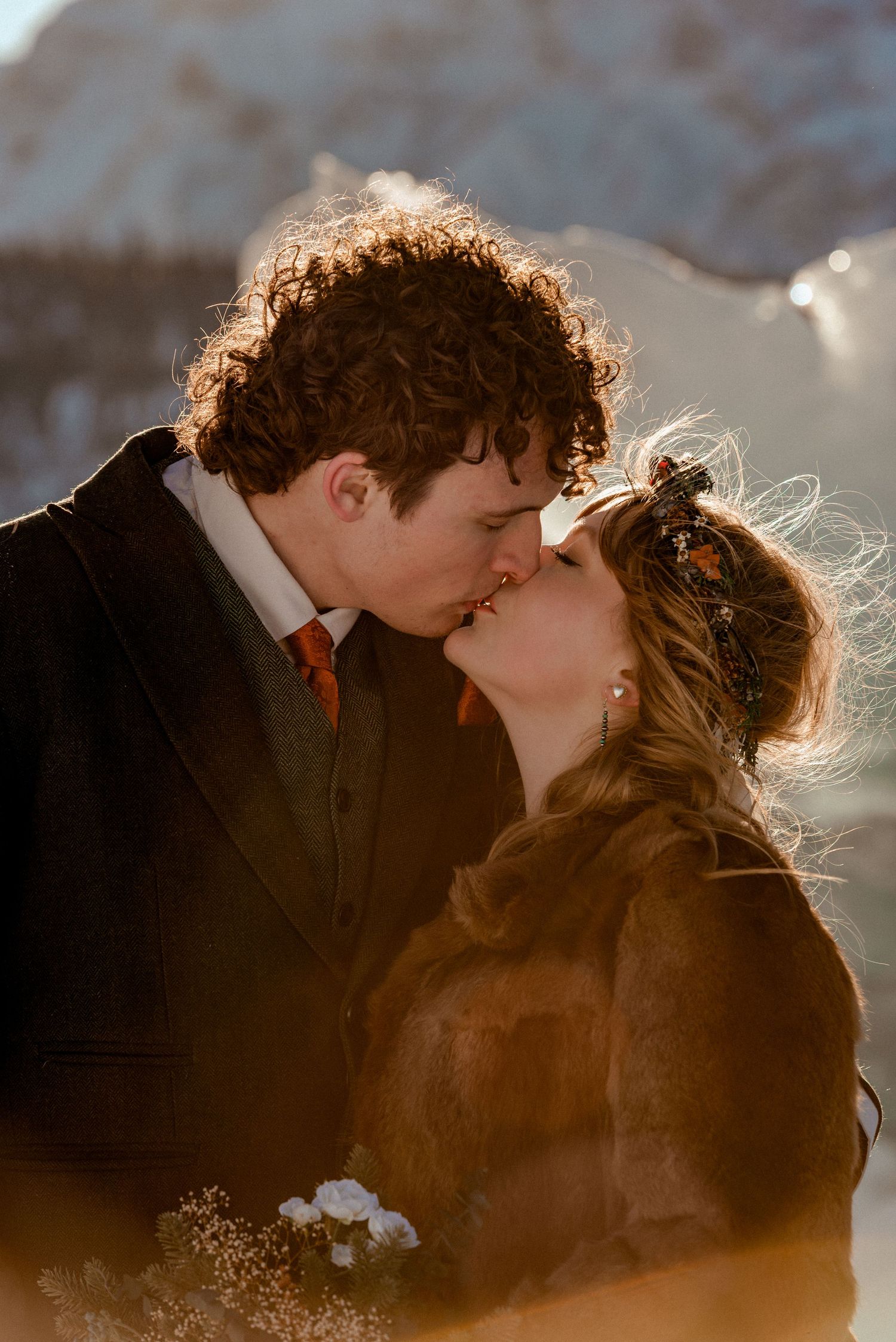 A couple shares a romantic kiss during their winter wedding, bathed in warm golden sunlight with snowy mountains behind them.