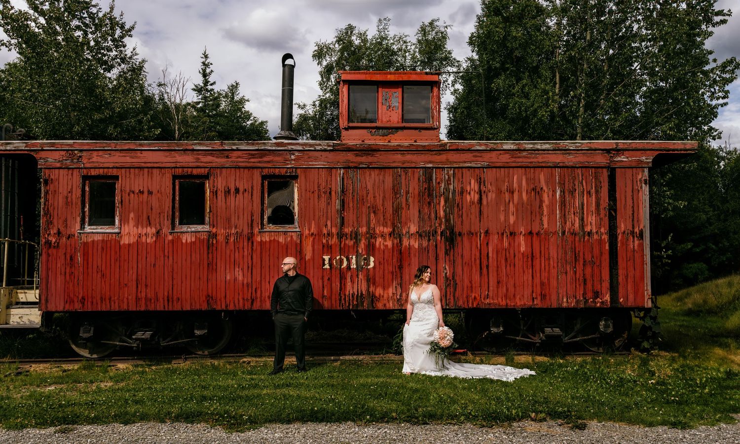 Bride and groom stand before a rustic red vintage train car outdoors on their wedding day.