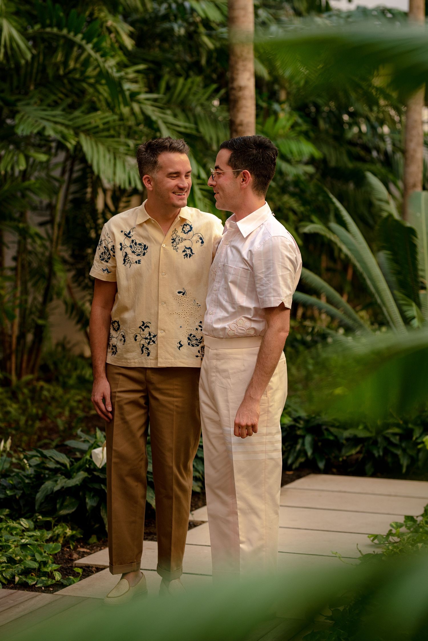 Two men in tropical attire smile at each other on a garden pathway surrounded by lush green plants.