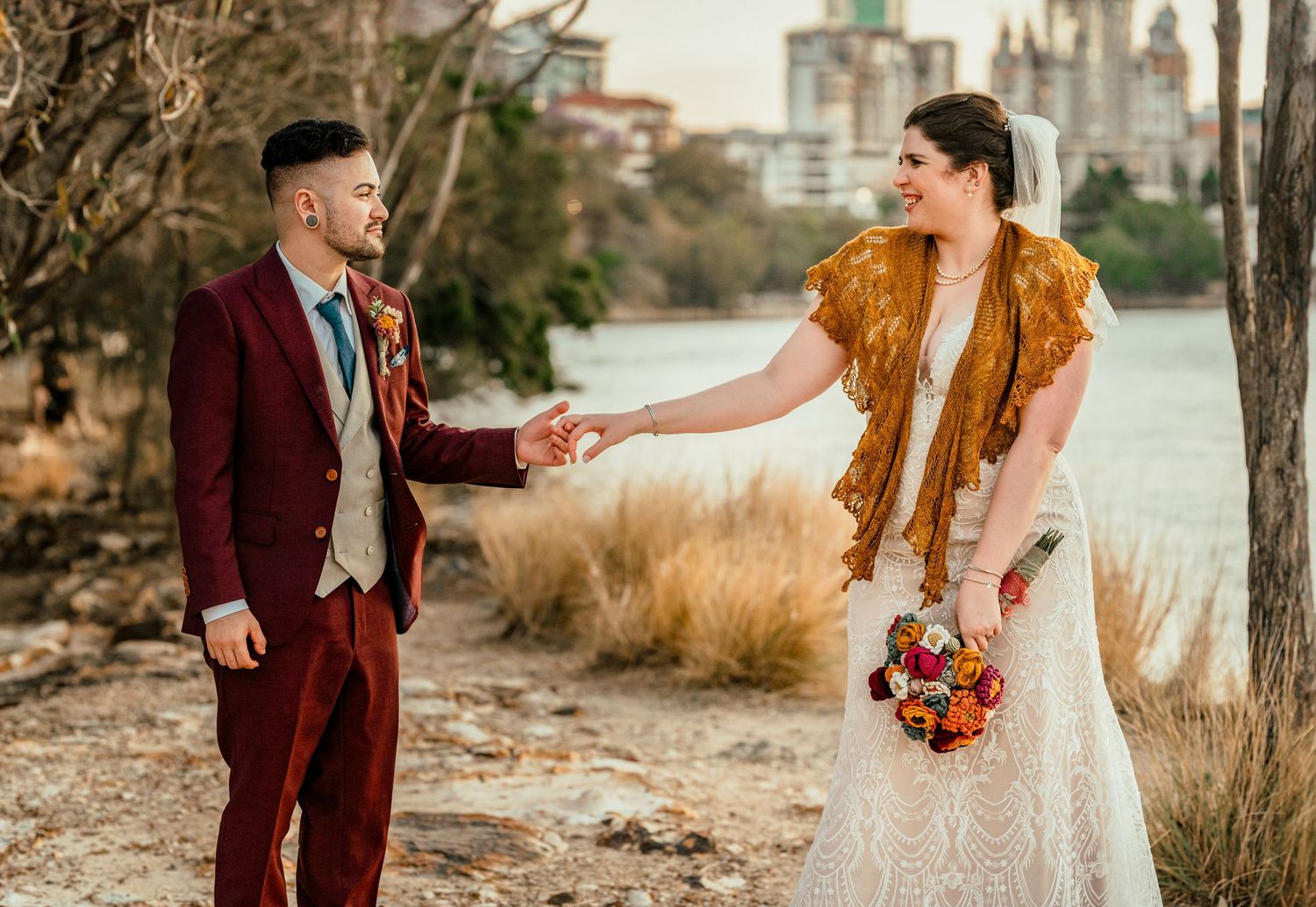 Bride in white lace gown with gold shawl holds groom's hand in burgundy suit by lakeside.