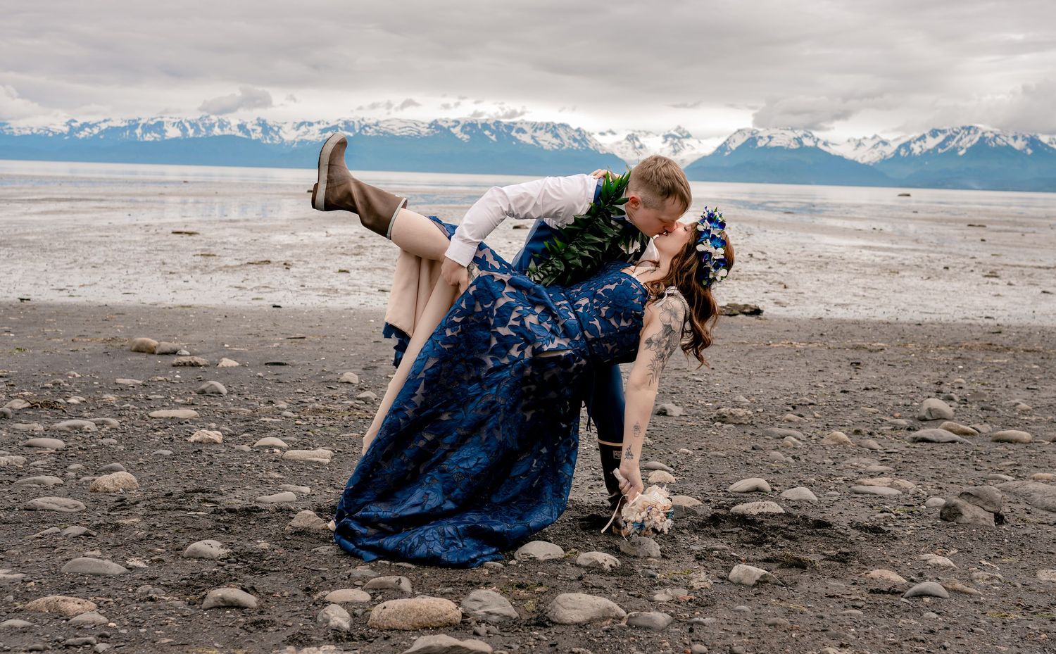 Couple sharing a romantic dip kiss on a rocky Alaskan beach with snow-capped mountains in the background.