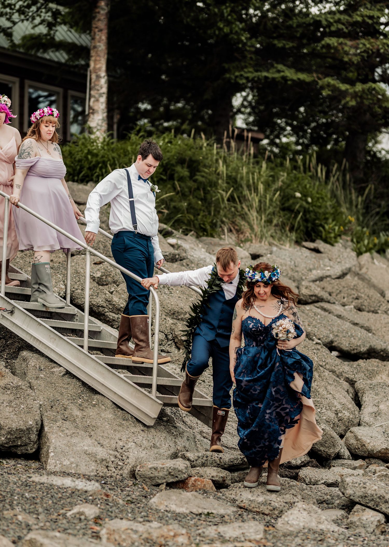 Wedding party navigating rocky shoreline, groom helping bride in navy floral gown while bridesmaids in pink descend metal stairs.