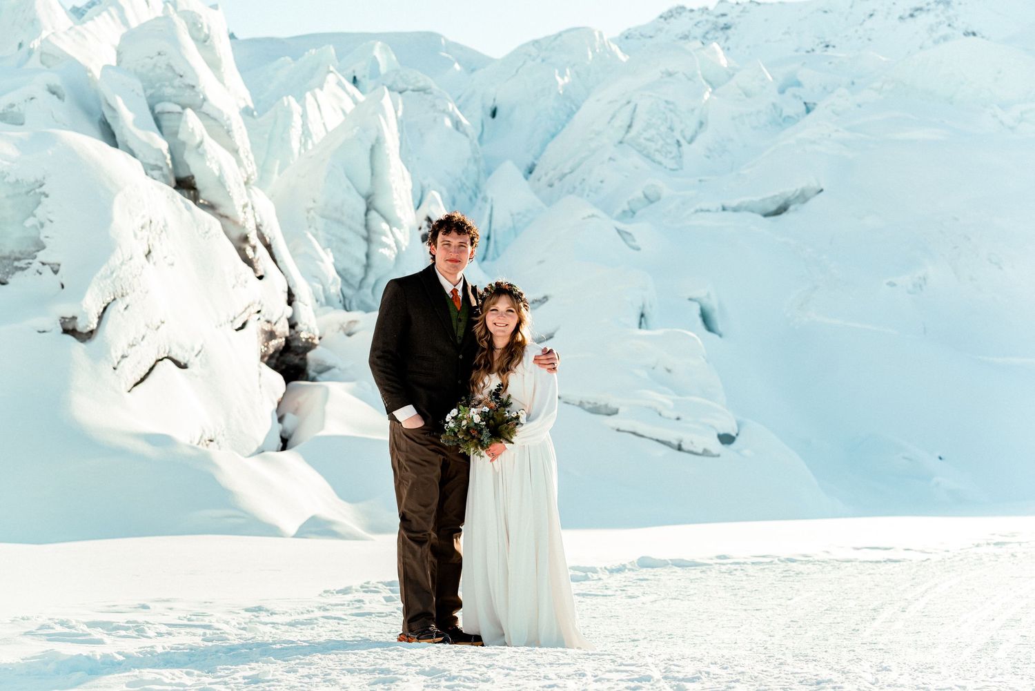 Couple in wedding attire posing on a snowy glacier, bride in white gown with floral crown, groom in dark suit.