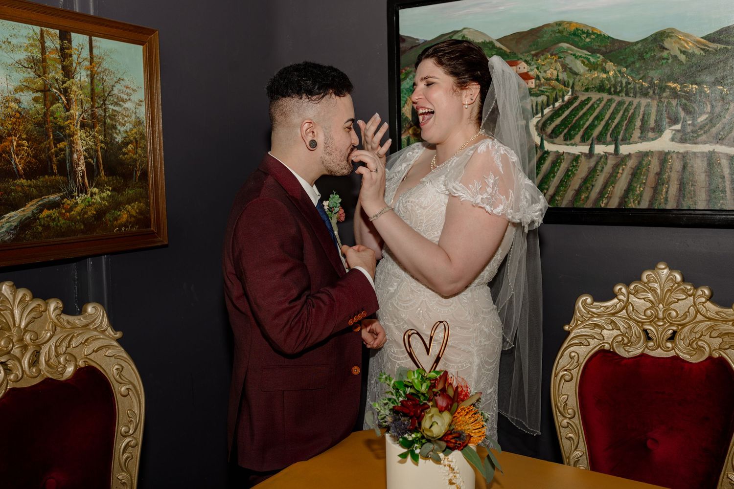 Bride and groom laughing together during wedding ceremony, bride in lace dress with veil, groom in burgundy suit.