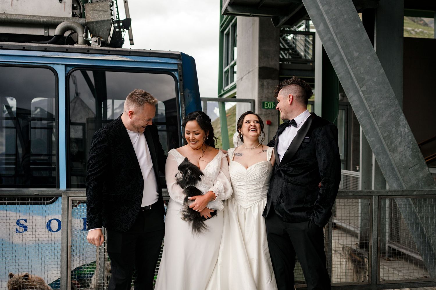Two brides in white gowns pose with guests and a black dog near a blue tram at an outdoor wedding venue.