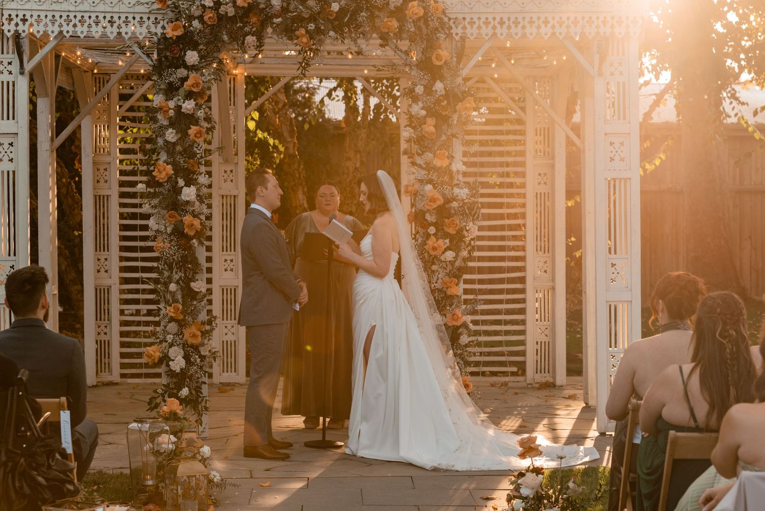 Couple exchanges vows at golden hour under floral arch at elegant outdoor wedding ceremony with sun flare casting a golden glow