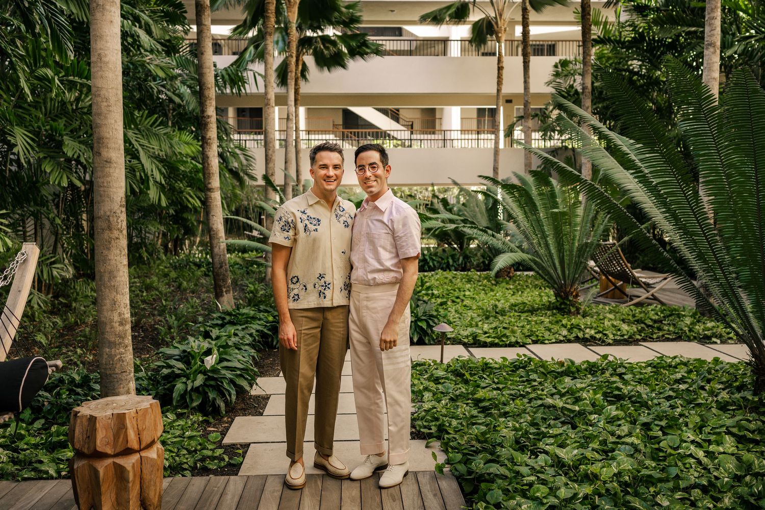 Two people posing together in a lush tropical garden atrium with green plants and a wooden walkway.