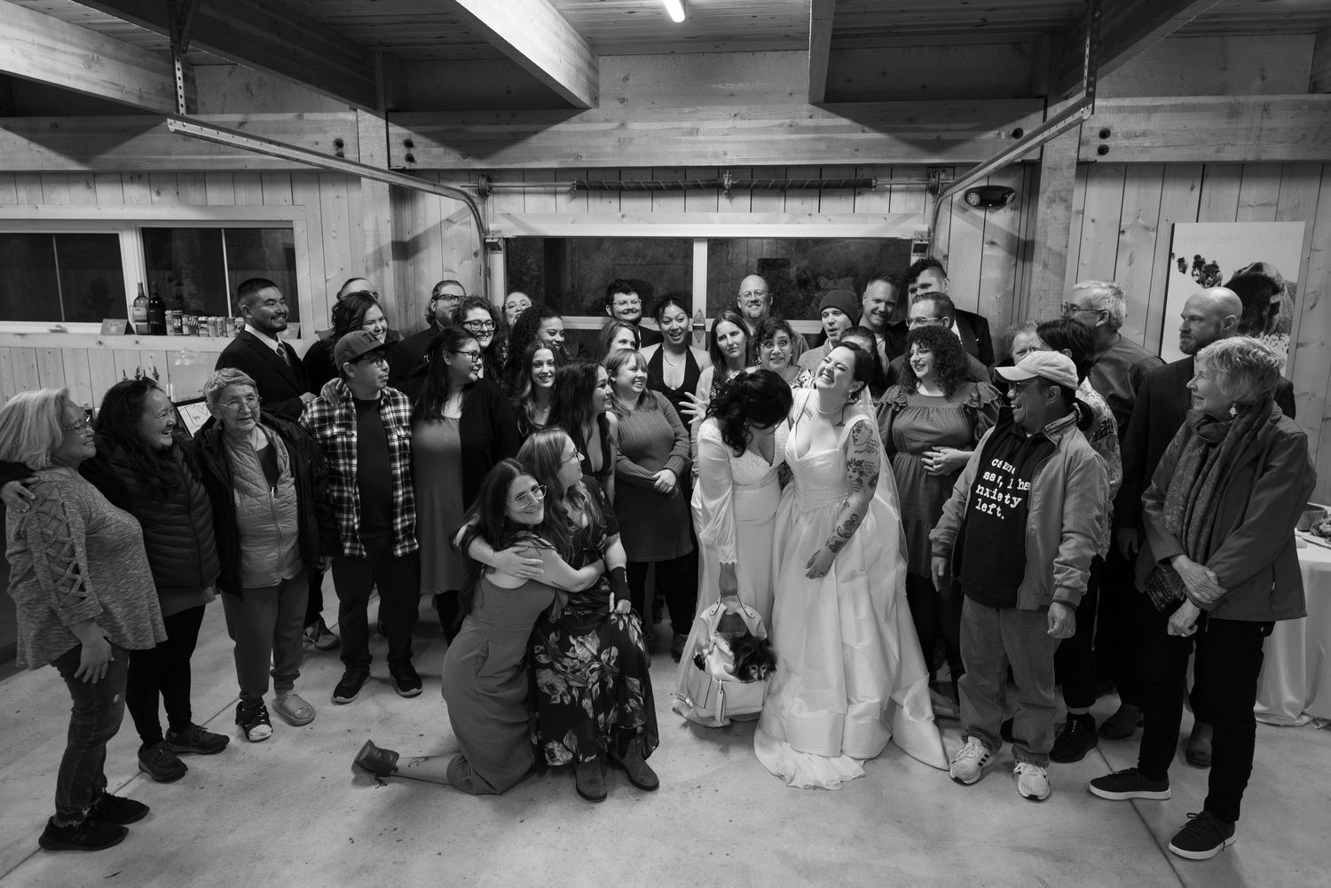 Black and white wedding photo of a bride and groom surrounded by a large group of smiling guests in a rustic venue.