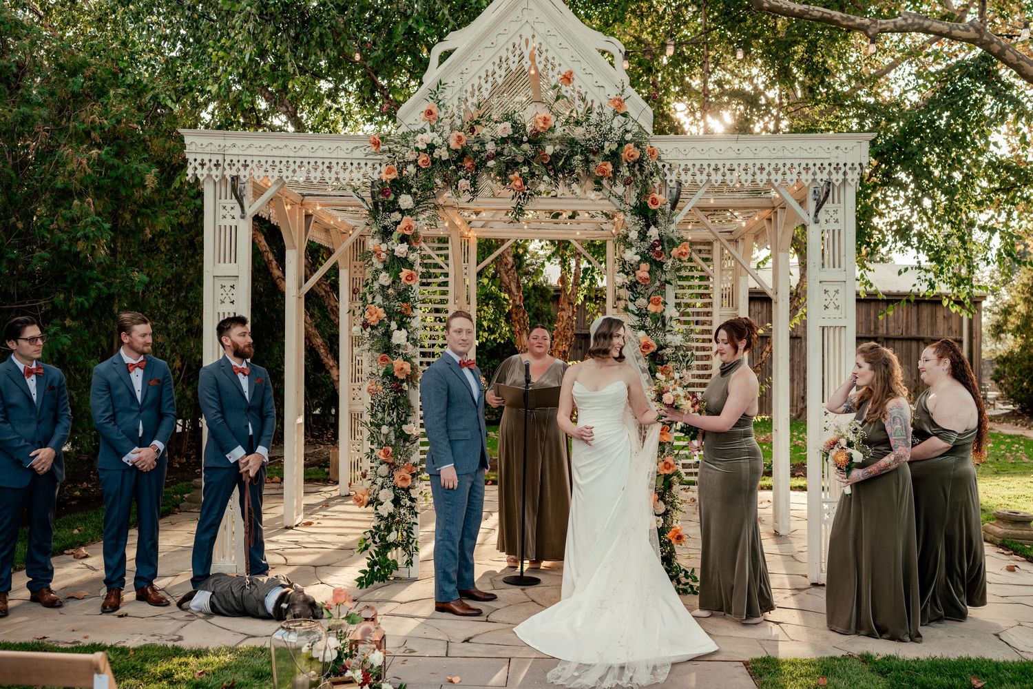 Bride and groom exchange vows under a floral gazebo surrounded by groomsmen and bridesmaids in olive dresses.