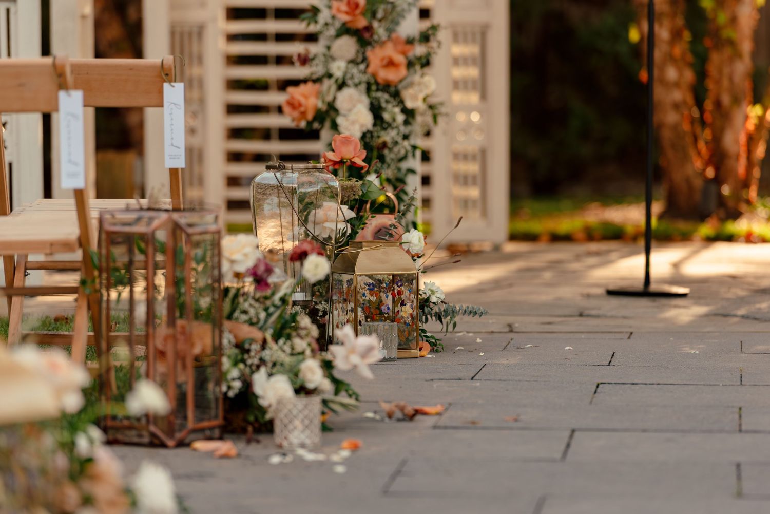 Elegant outdoor wedding ceremony aisle with lanterns, floral arrangements, and wooden chairs on stone pavers.