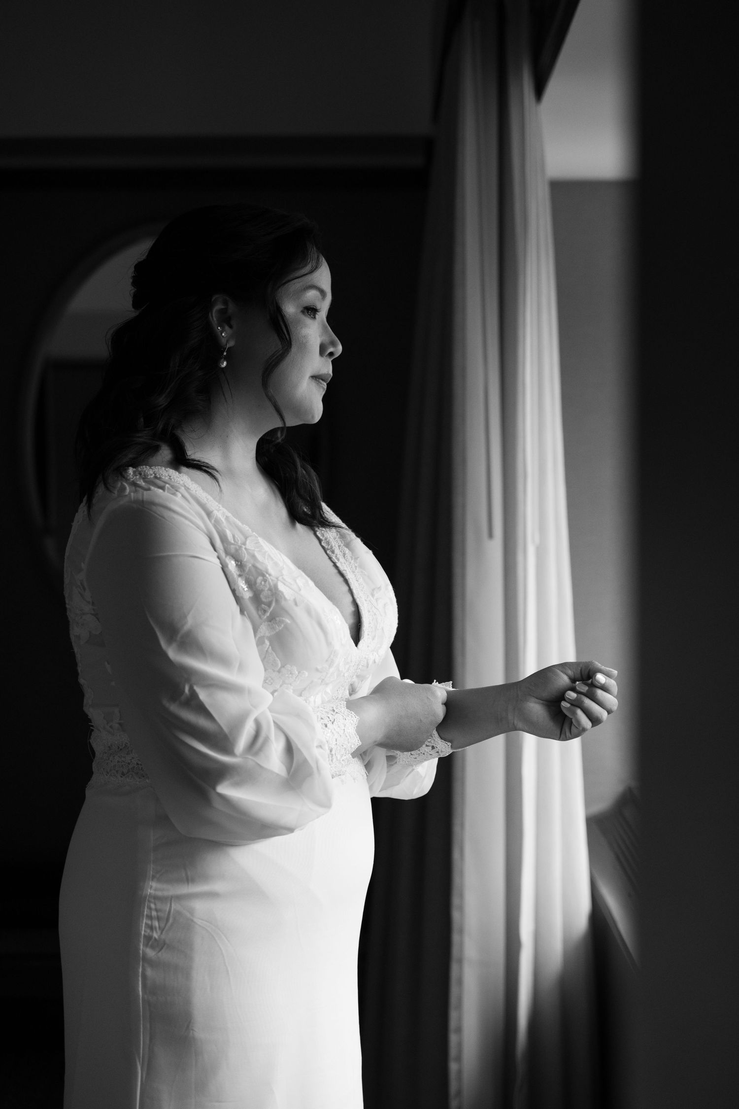 Bride in white lace wedding dress standing by window in black and white portrait photo.