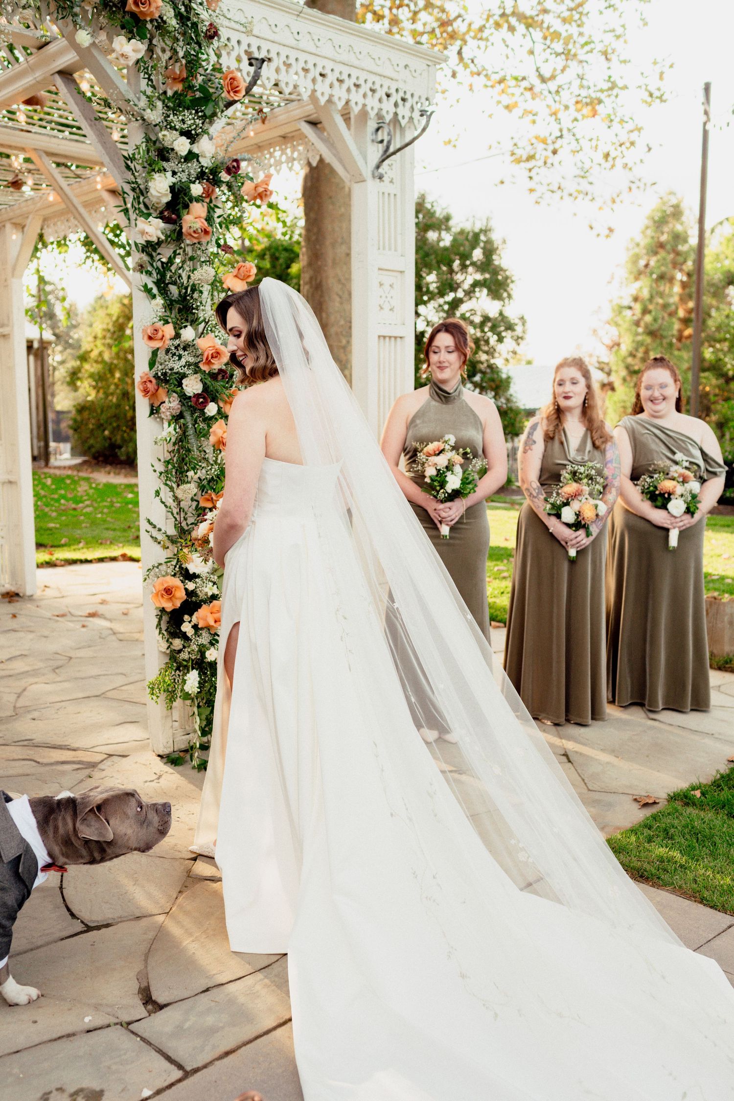 Bride in white gown and veil stands at floral arch with bridesmaids in olive dresses holding bouquets at the altar in Summer
