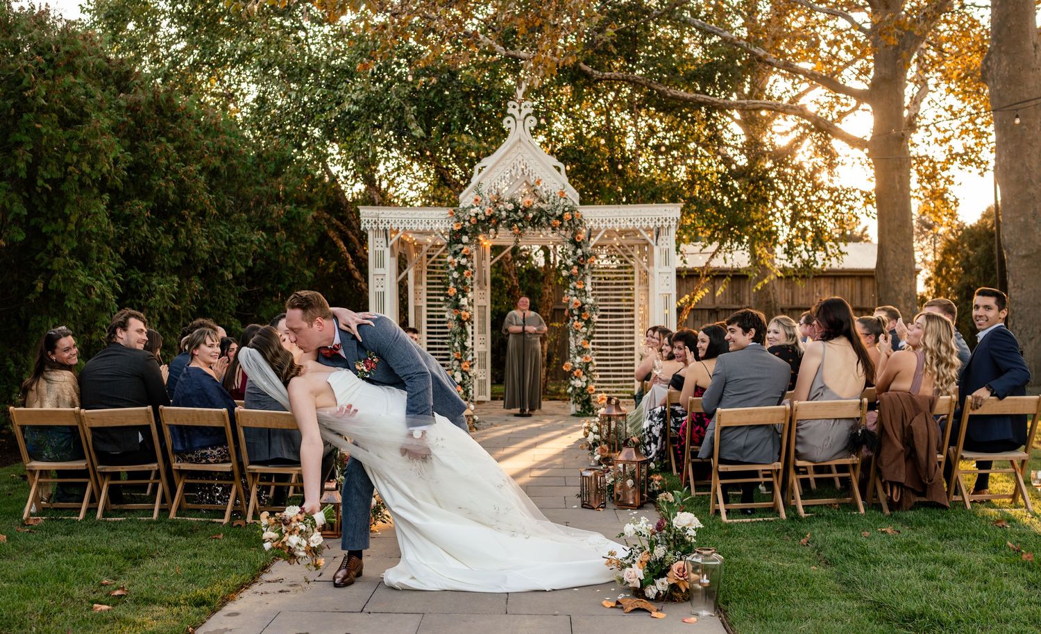 Bride and groom share romantic dip kiss at outdoor garden wedding ceremony with white gazebo and seated guests at golden hour.