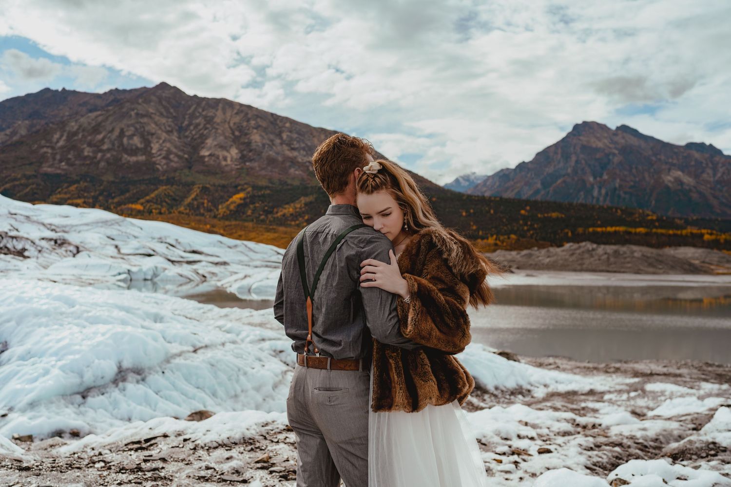 Couple wrapped in a blanket stands together against dramatic mountain and glacier landscape at sunset.