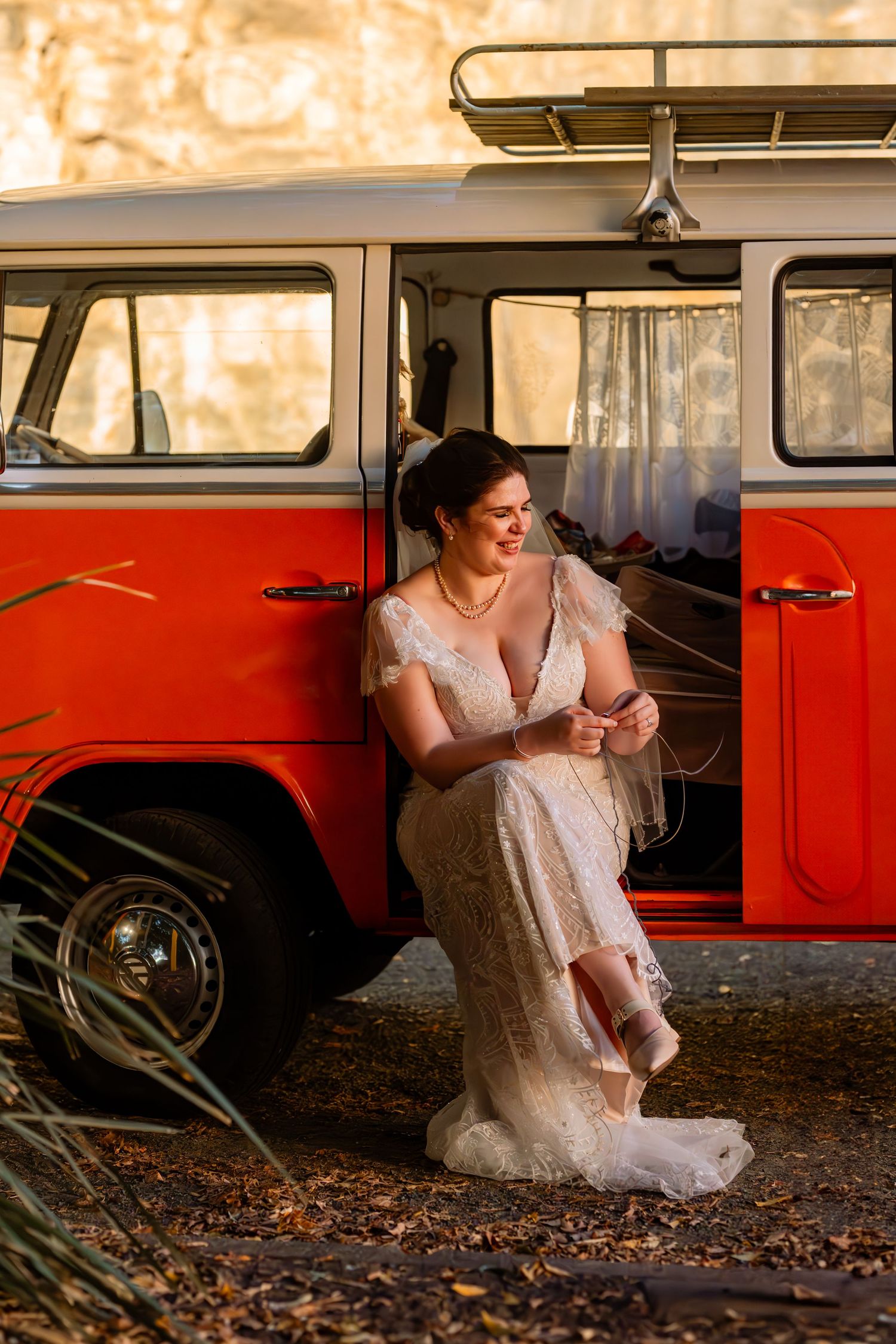 Bride in lace wedding dress leaning against a vintage red VW van, adjusting her shoe in golden hour light.