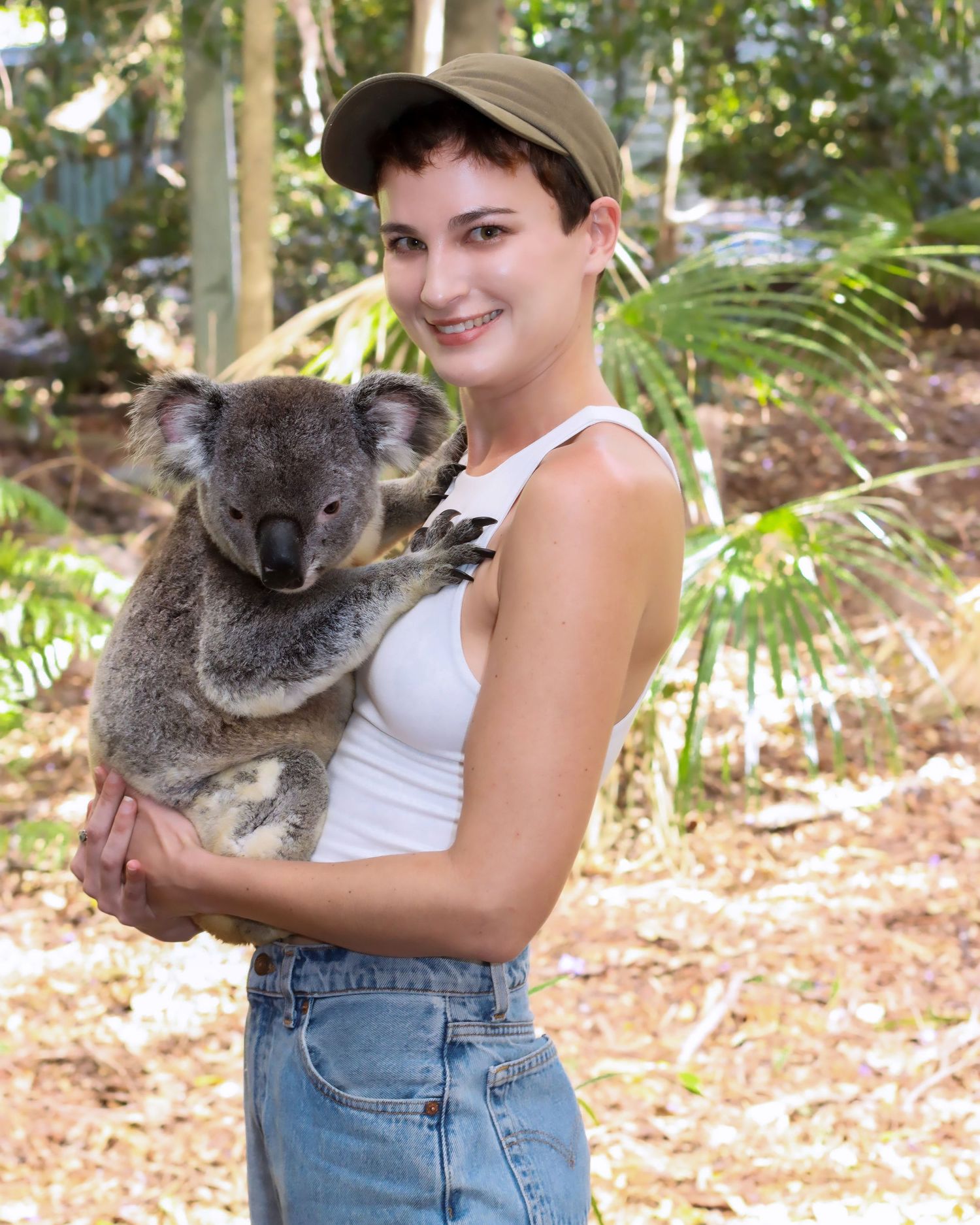 Young woman with short hair holding a koala bear in an outdoor Australian wildlife sanctuary setting.