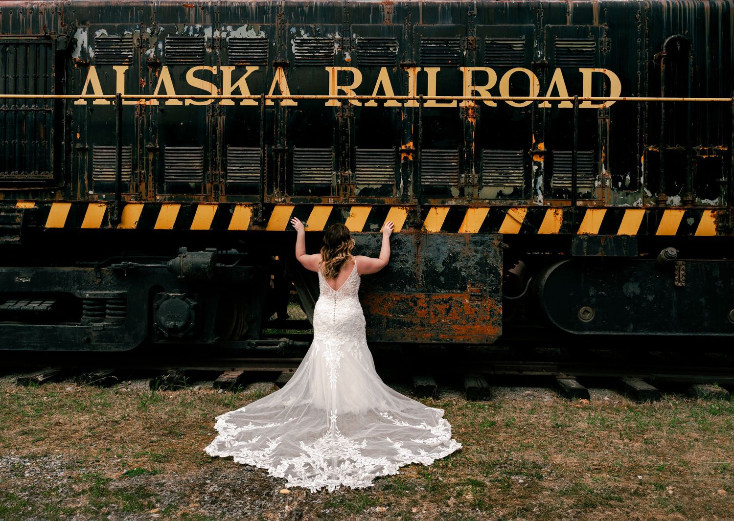 Bride in lace wedding gown poses before vintage Alaska Railroad train car on her wedding day.