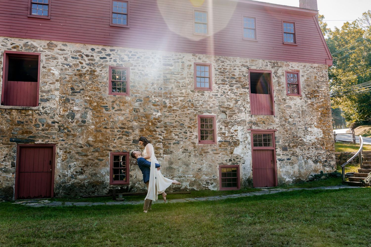 Couple dancing in front of a rustic stone building with red-trimmed windows and doors on a sunny day.