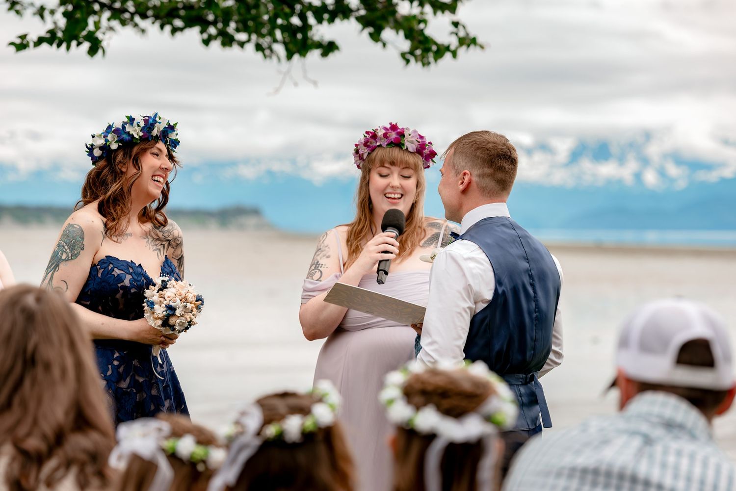 Outdoor beach wedding ceremony with flower crown bride laughing as officiant speaks into microphone near Alaska mountains.