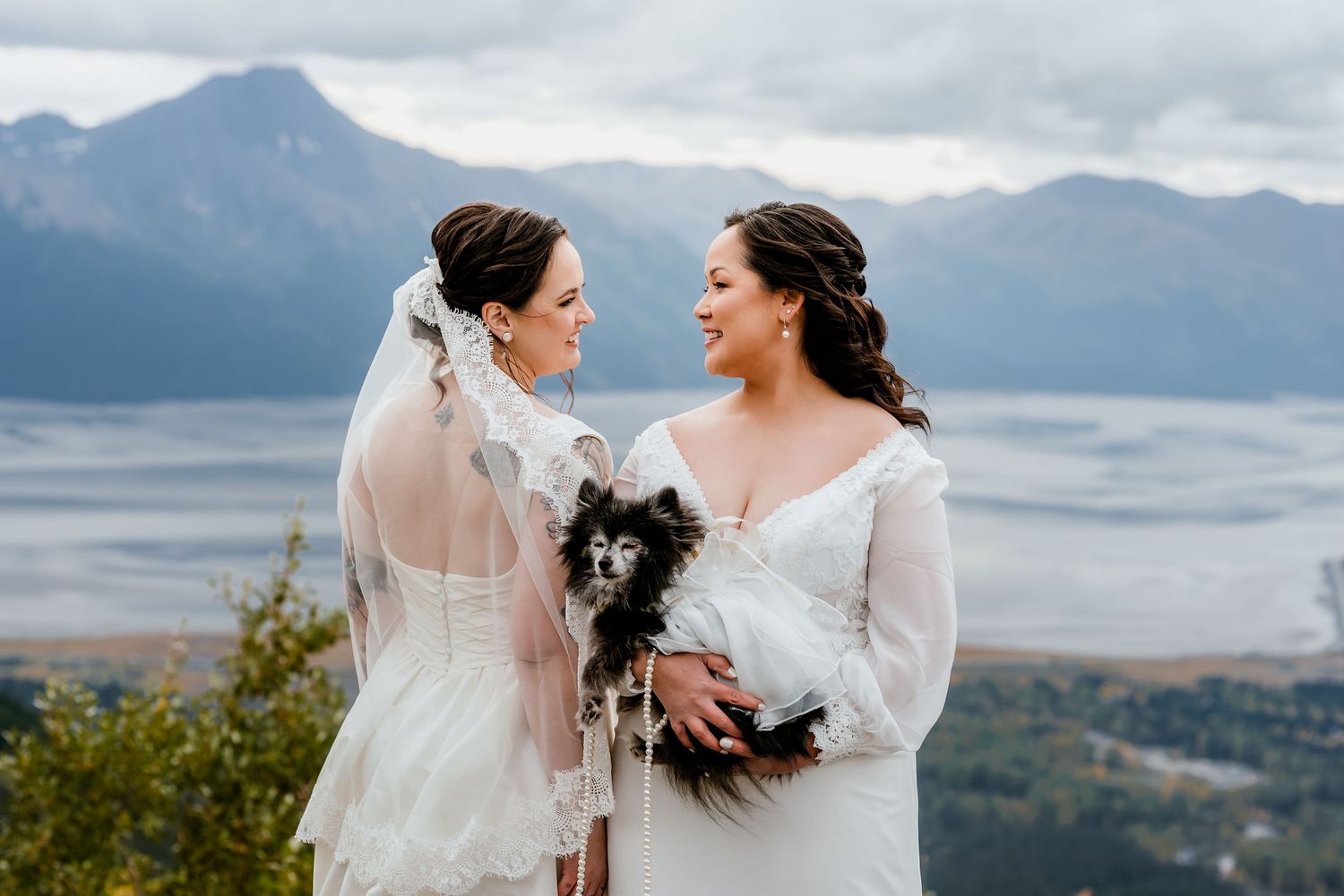 Two brides in white gowns share a joyful moment holding a small dog with mountain views behind them.