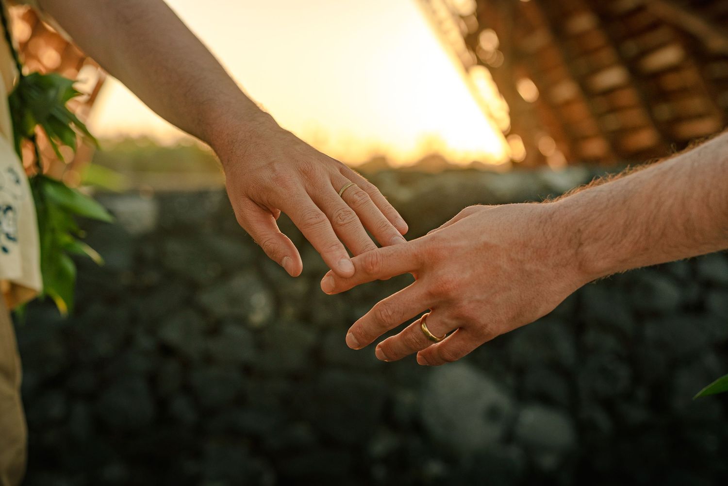 Two hands gently reaching toward each other at sunset, one wearing a gold ring, with warm golden light in the background.