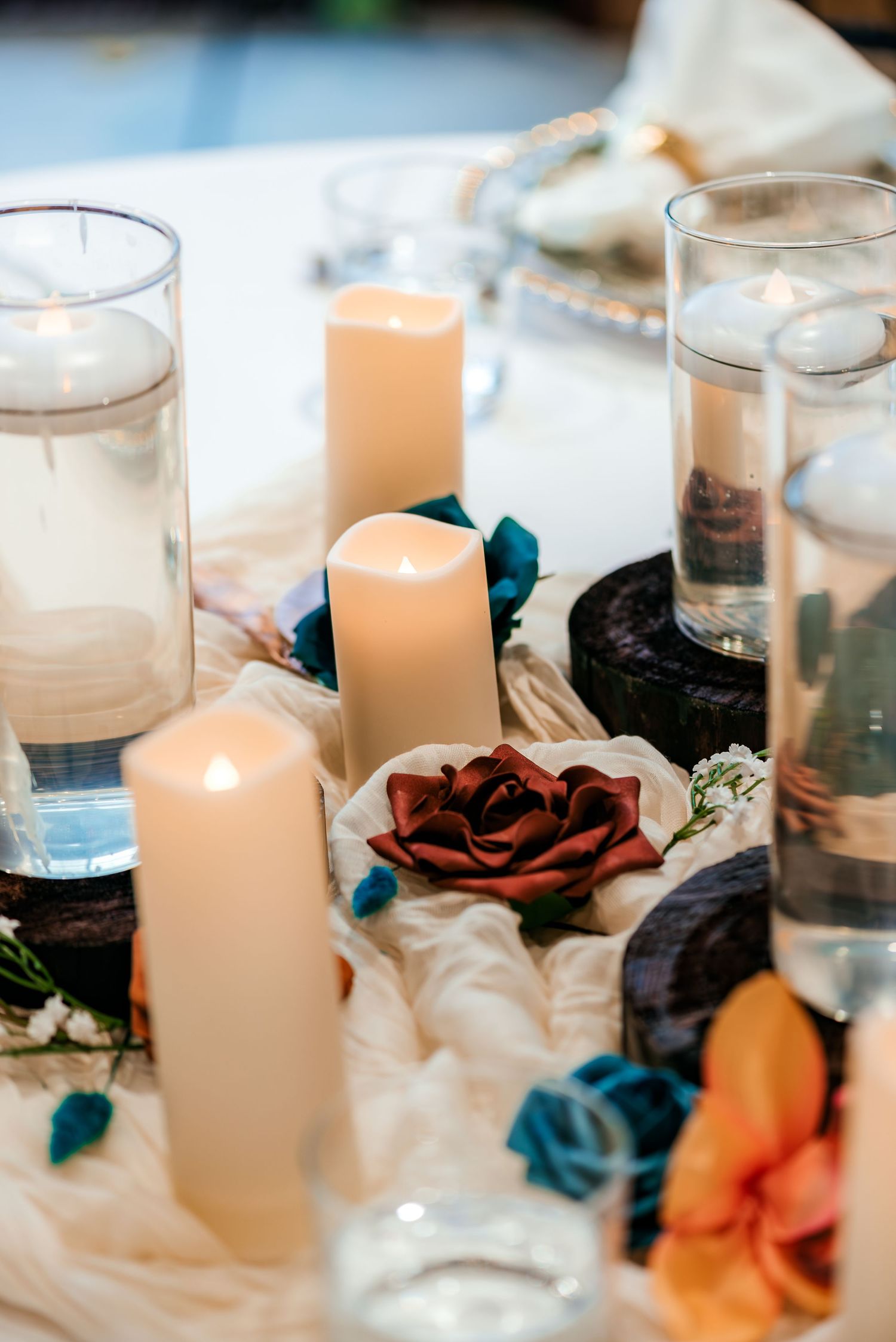 Elegant wedding table centerpiece with white pillar candles, teal and burgundy roses, and dark wood accents on white fabric.