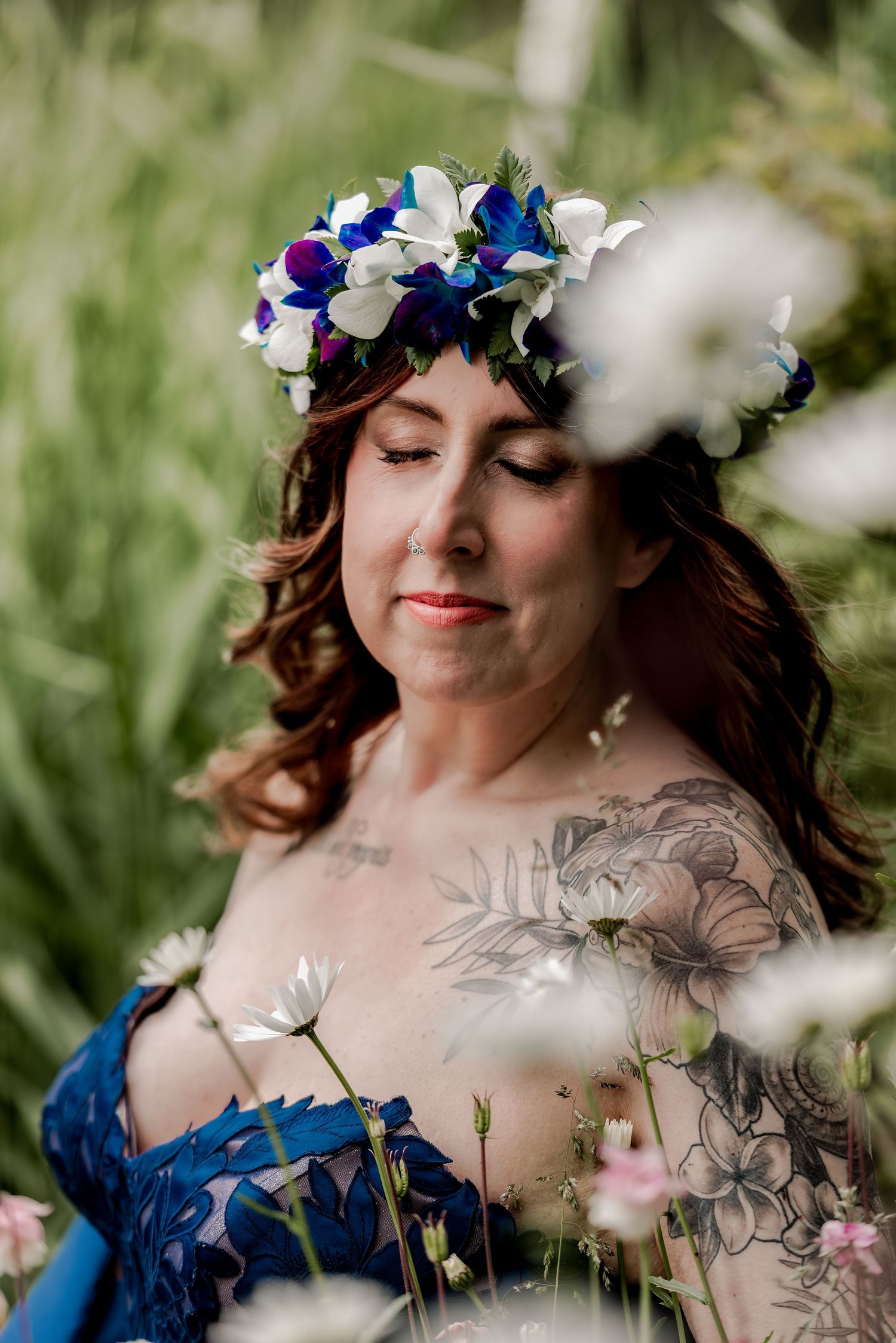 Woman with floral crown and tattoos poses among wildflowers in a blue lace dress, eyes closed peacefully.