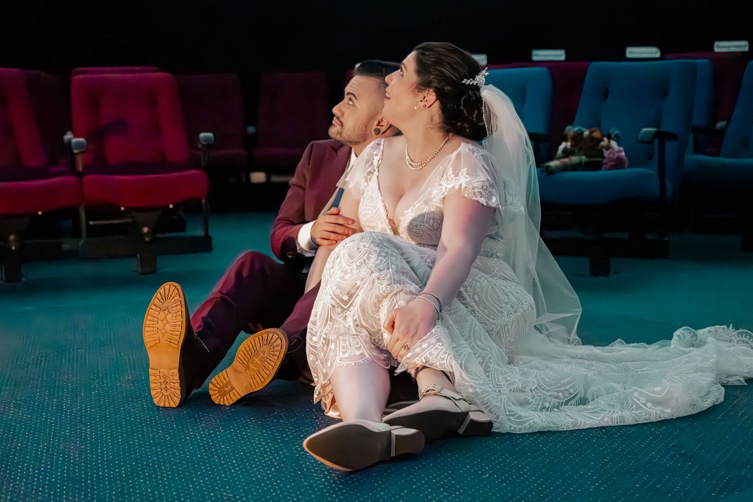 Bride and groom in wedding attire sit on floor of theater, sharing a joyful moment among red and purple seats.