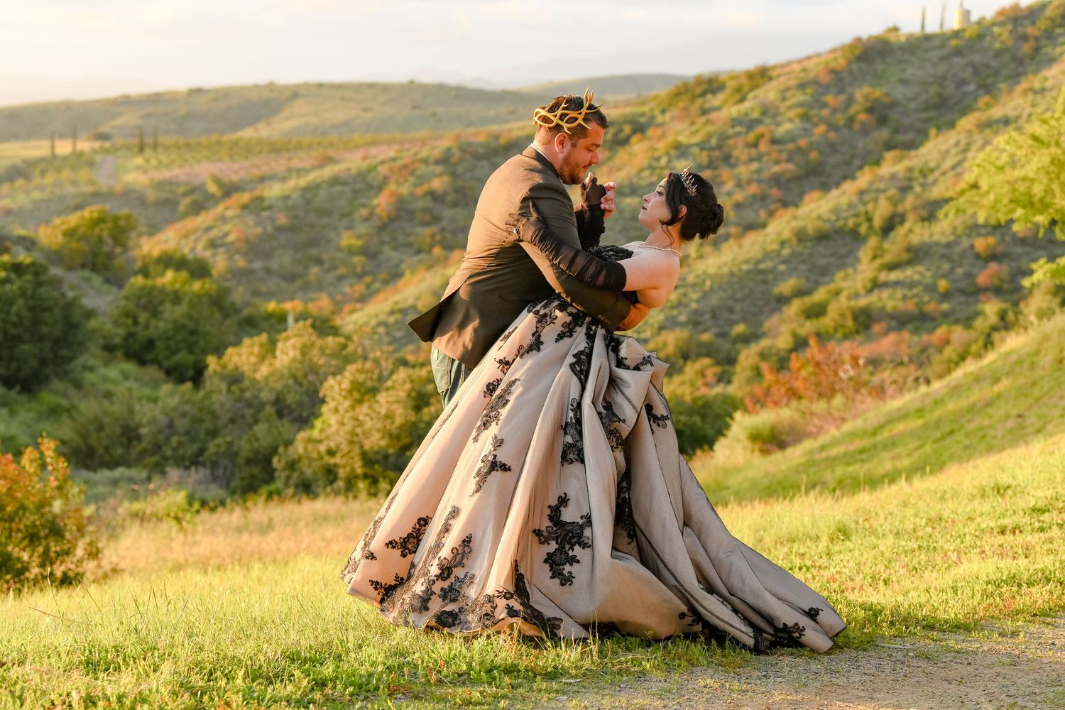 Couple in wedding attire embrace dramatically on a hilltop at sunset.