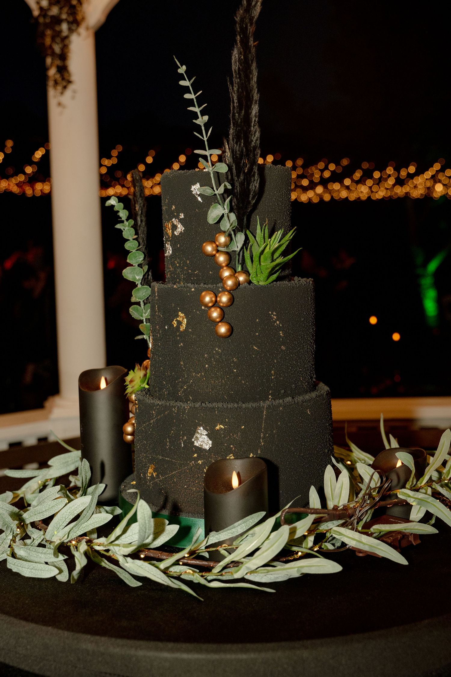 Elegant three-tier black wedding cake with gold accents, greenery, and dark candles on a table at night.