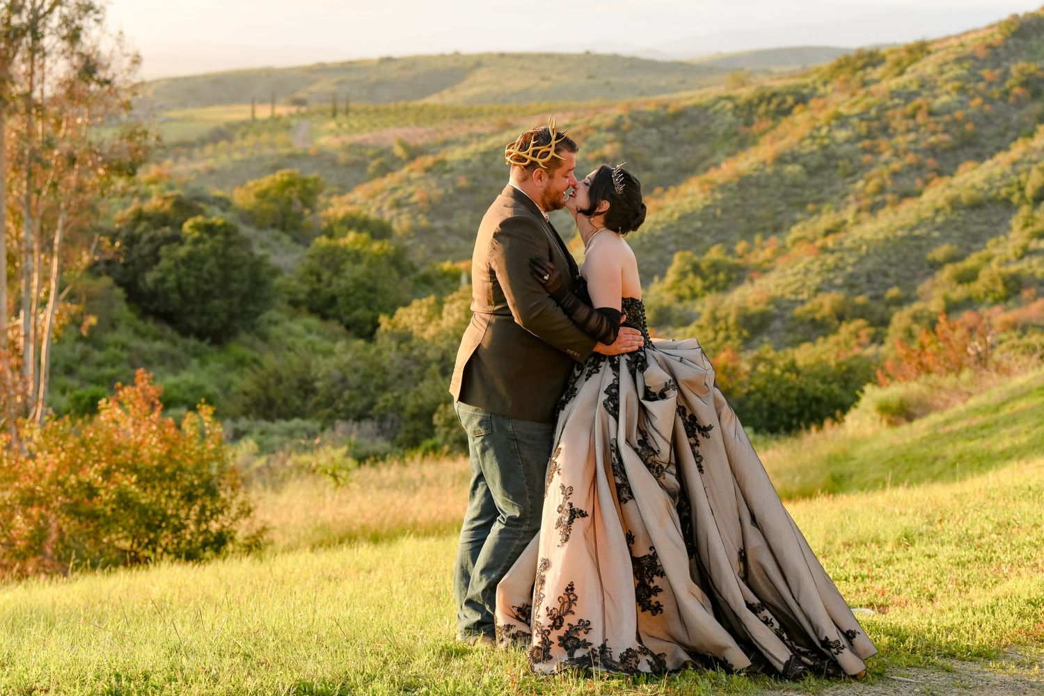 Couple sharing a romantic kiss in a scenic green hillside, bride wearing an elegant floral gown at golden hour.