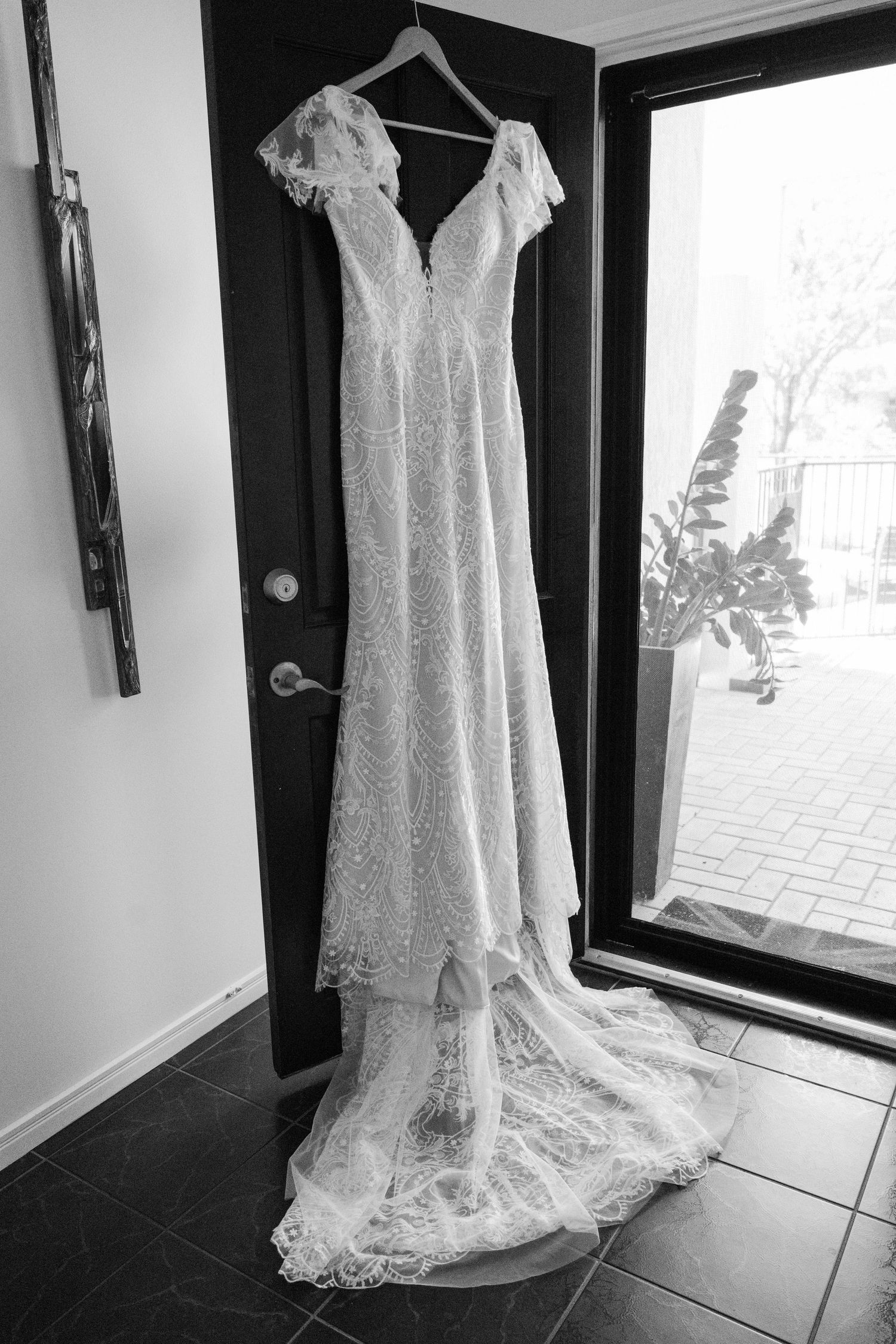 Black and white photo of an elegant lace wedding dress hanging on a door with a train pooling on the floor.