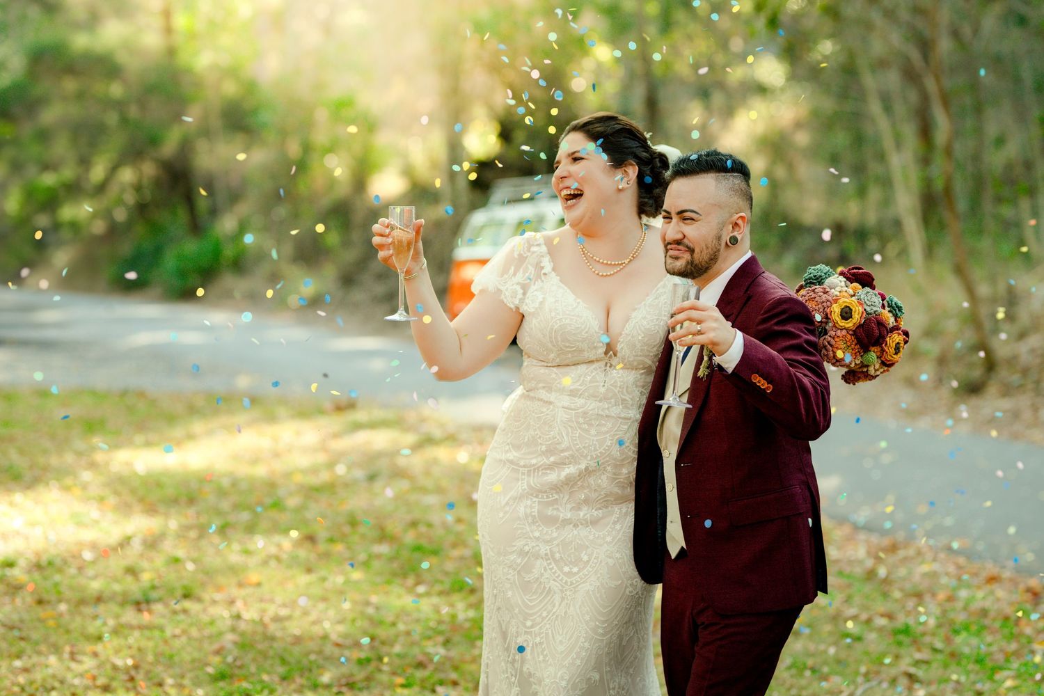 Joyful bride and groom laughing outdoors, confetti flying, groom holding sunflower bouquet in burgundy suit.