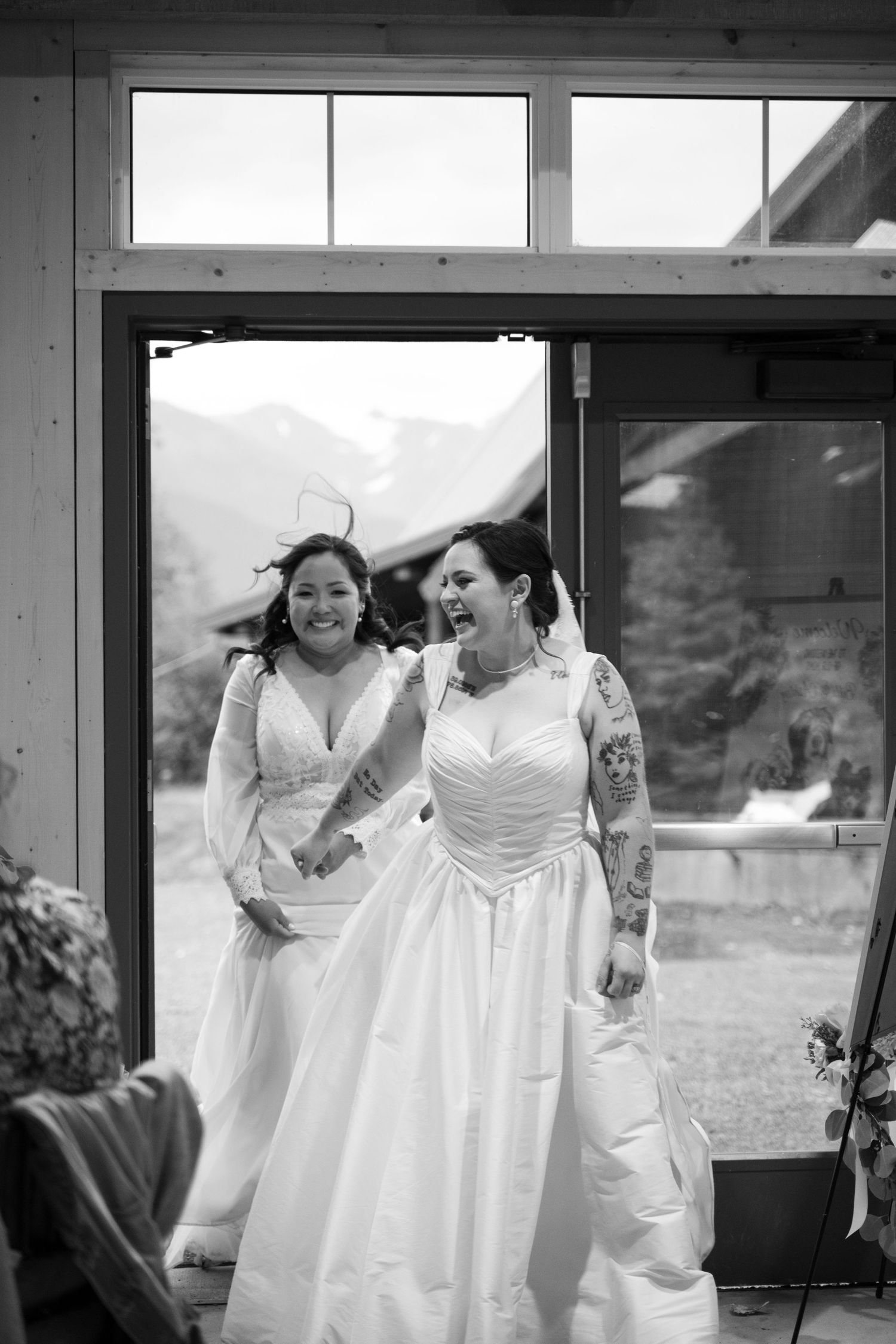 Two brides in white wedding gowns laugh joyfully as they enter a venue, with mountain scenery visible through the doorway.