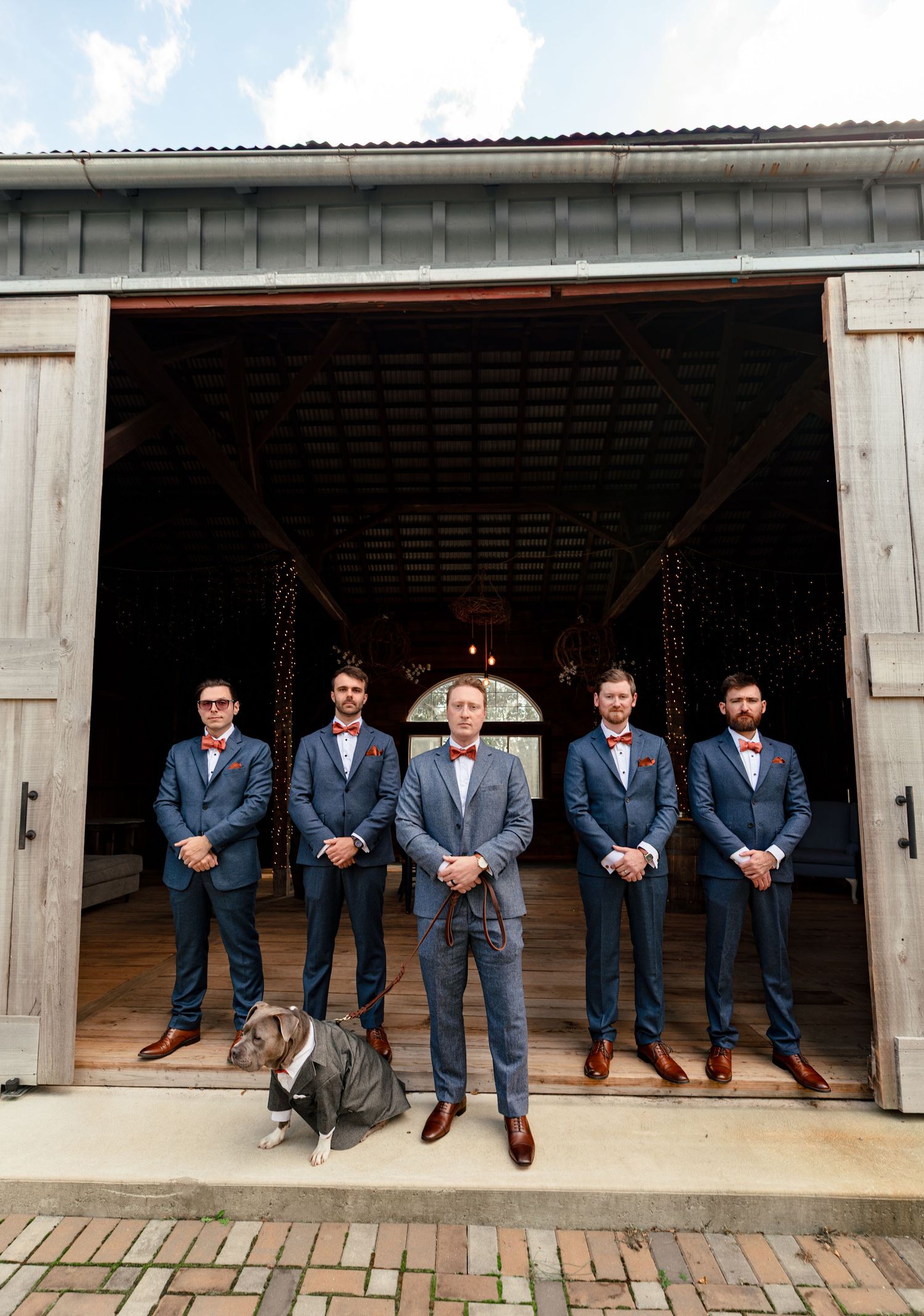 Five groomsmen in navy suits with red bow ties pose with a Pitbull outside rustic barn wedding venue.