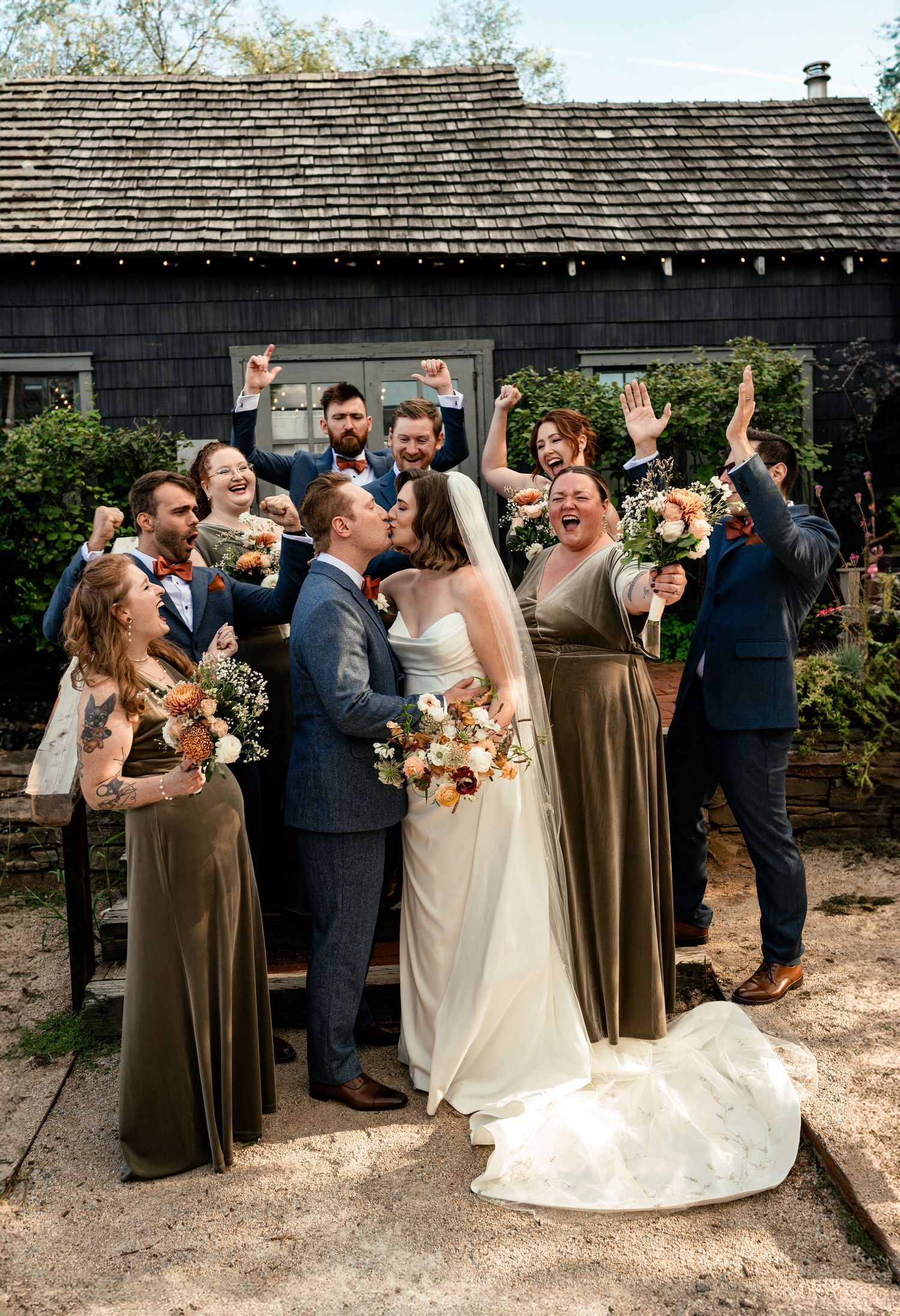 Wedding party celebrating as bride and groom kiss outdoors, bridesmaids in olive dresses, groomsmen in navy suits.